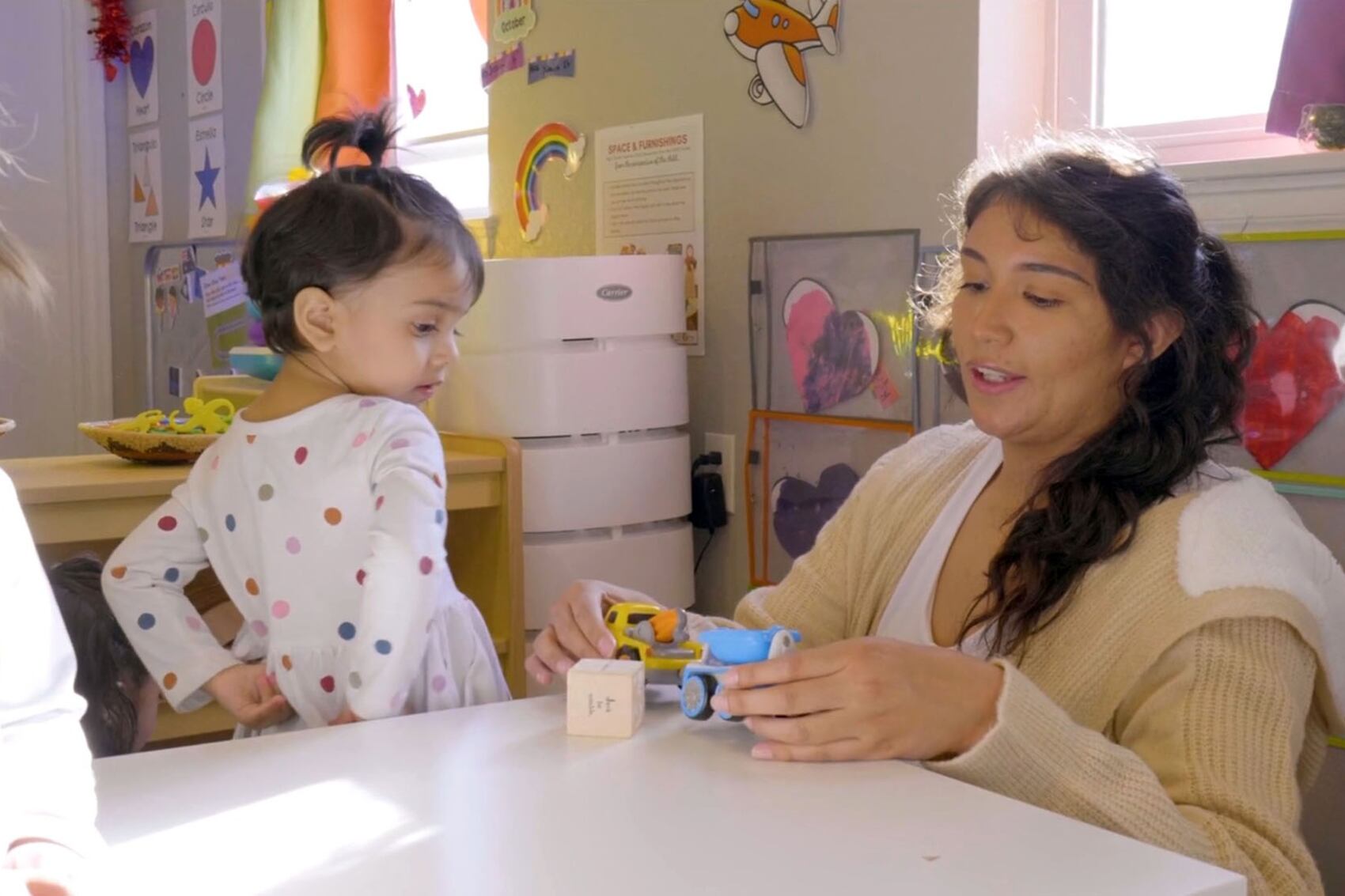 A teacher with dark hair plays with toys at a table with a young child in an early childhood education classroom.