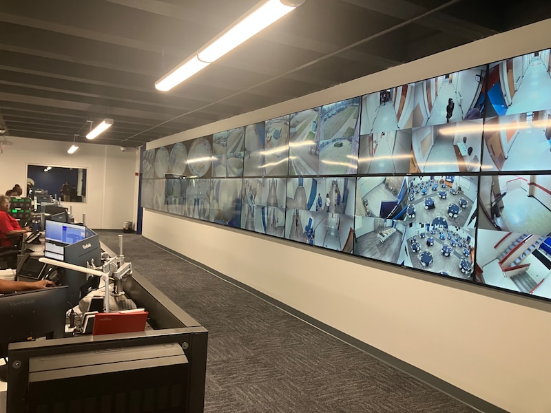 A wall lined with 18 televisions shows live camera footage of a building hallway, cafeteria, and different doorways. In front of the TVs are a row of desktop computers, with people typing.