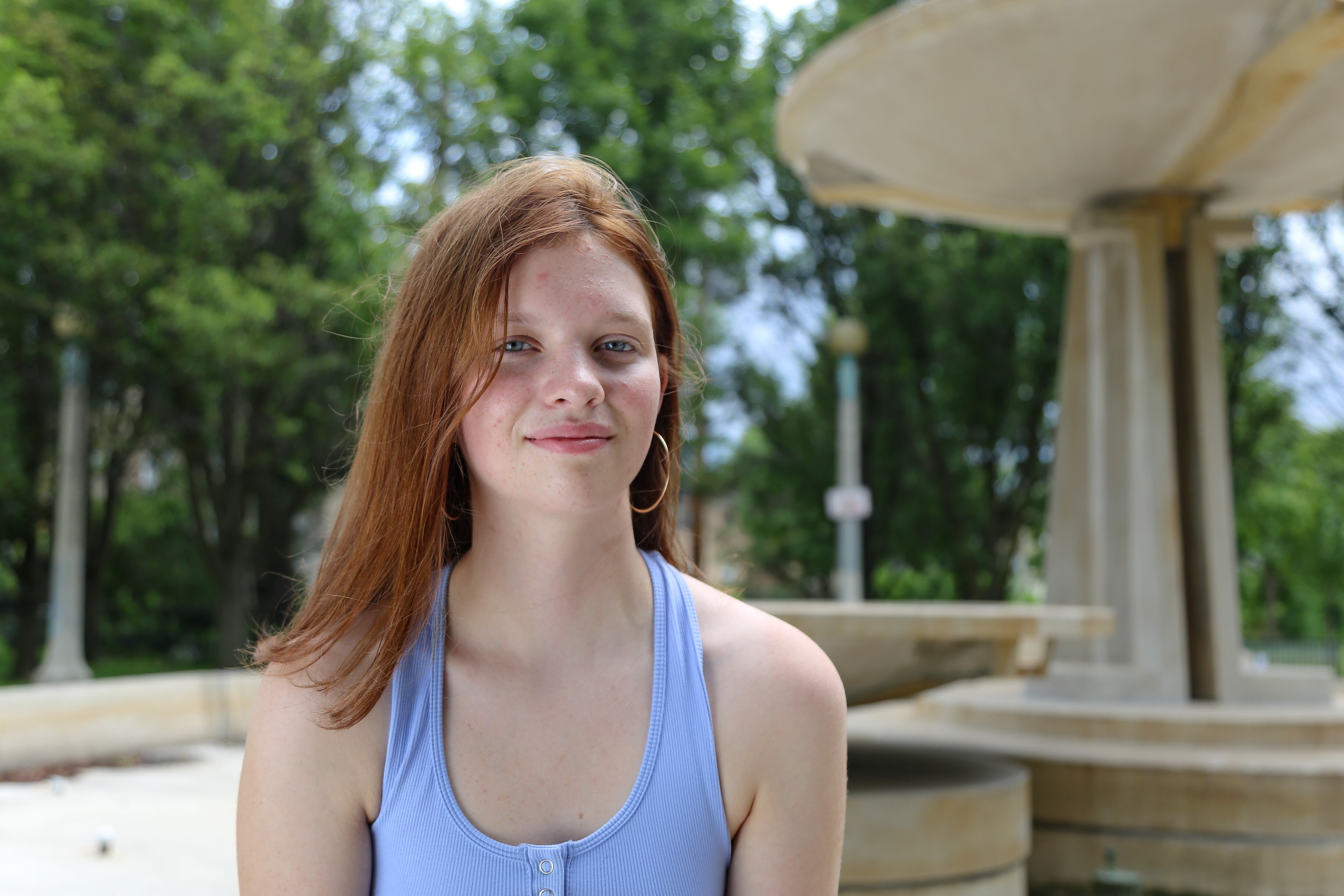 A young woman with auburn hair poses for a portrait next to a fountain in a public park.