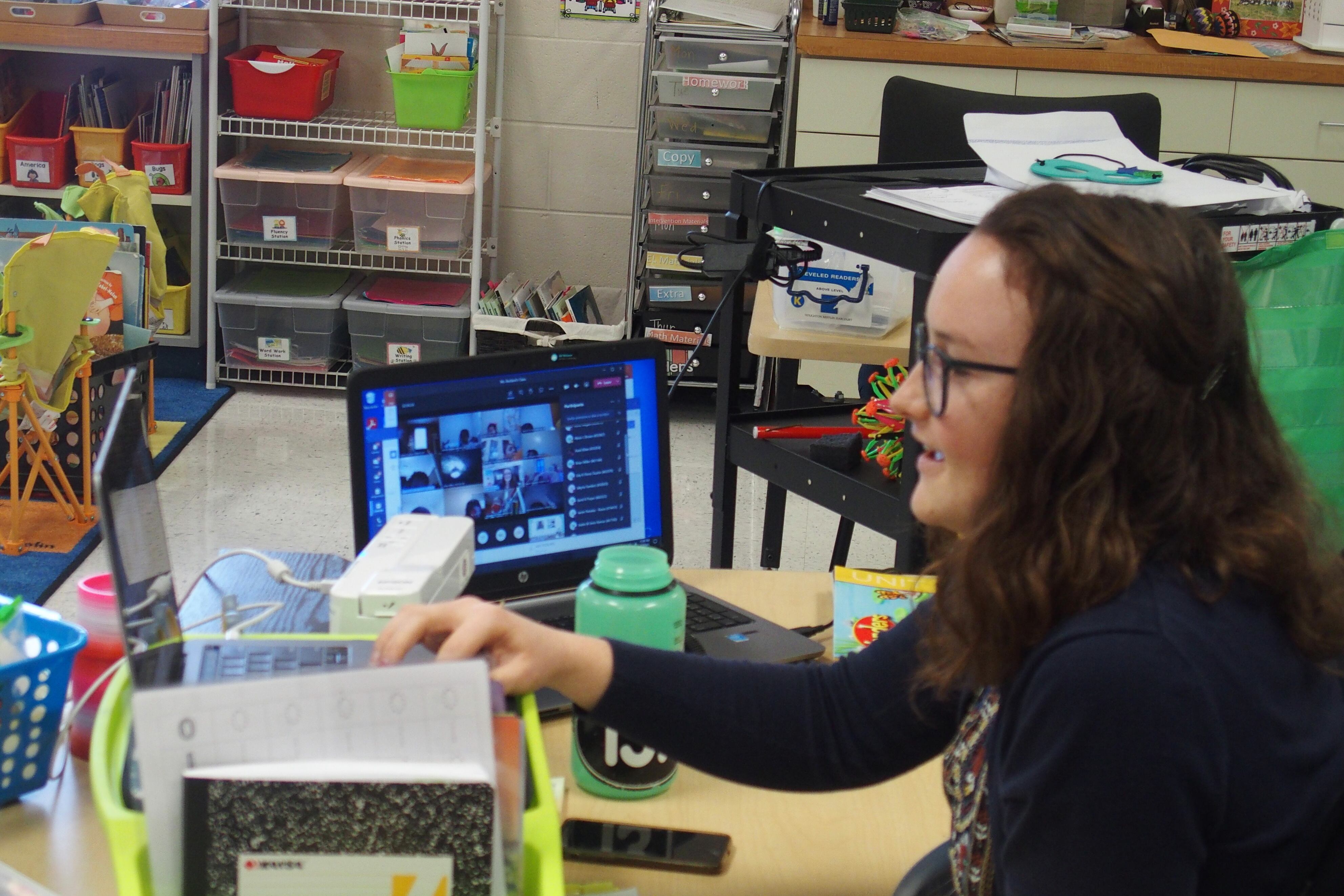 A teacher has two laptops set up at her classroom desk so she can instruct and see her students on videoconference.