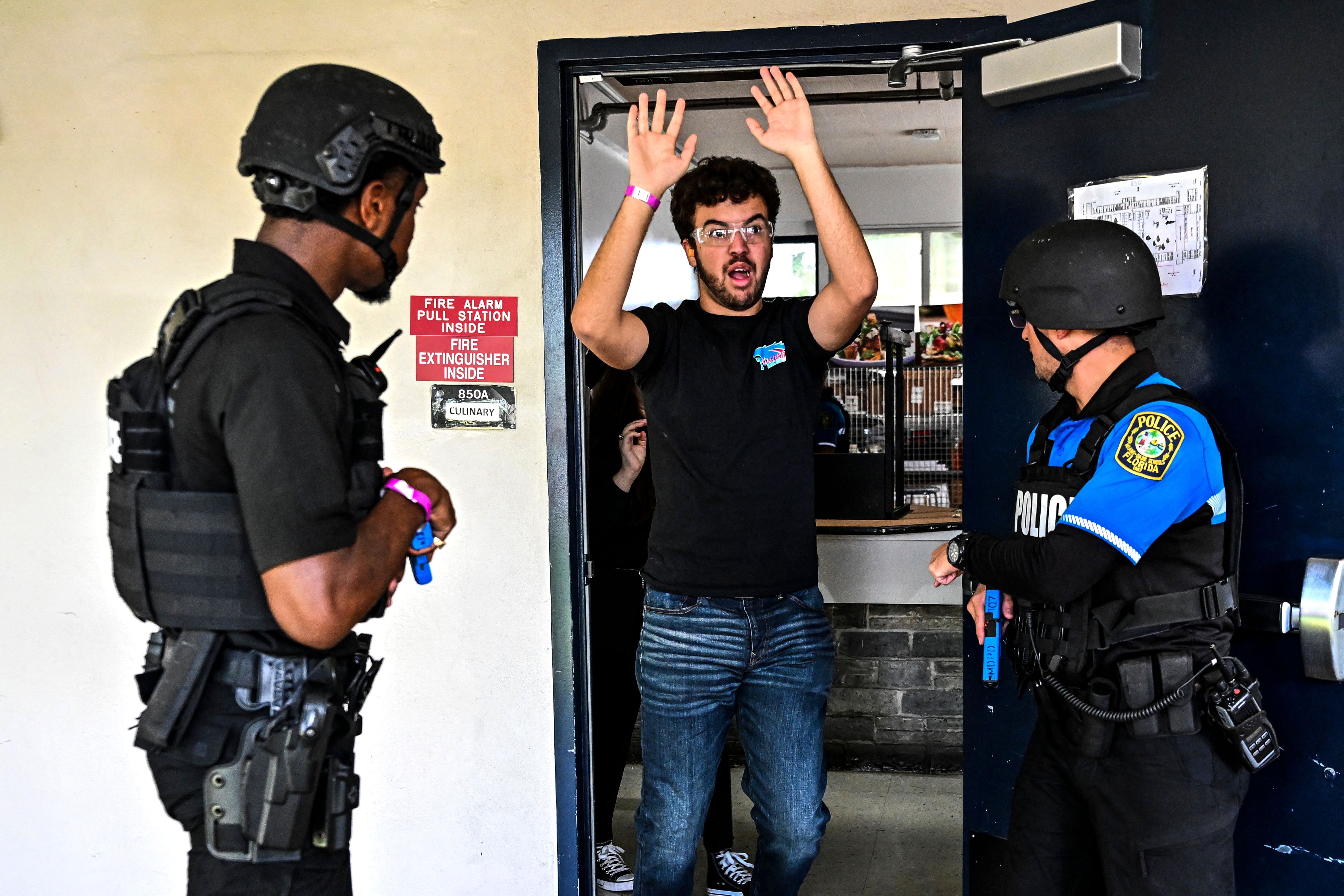 Two police officers have their guns as a young man walks out of a door with his hands up.