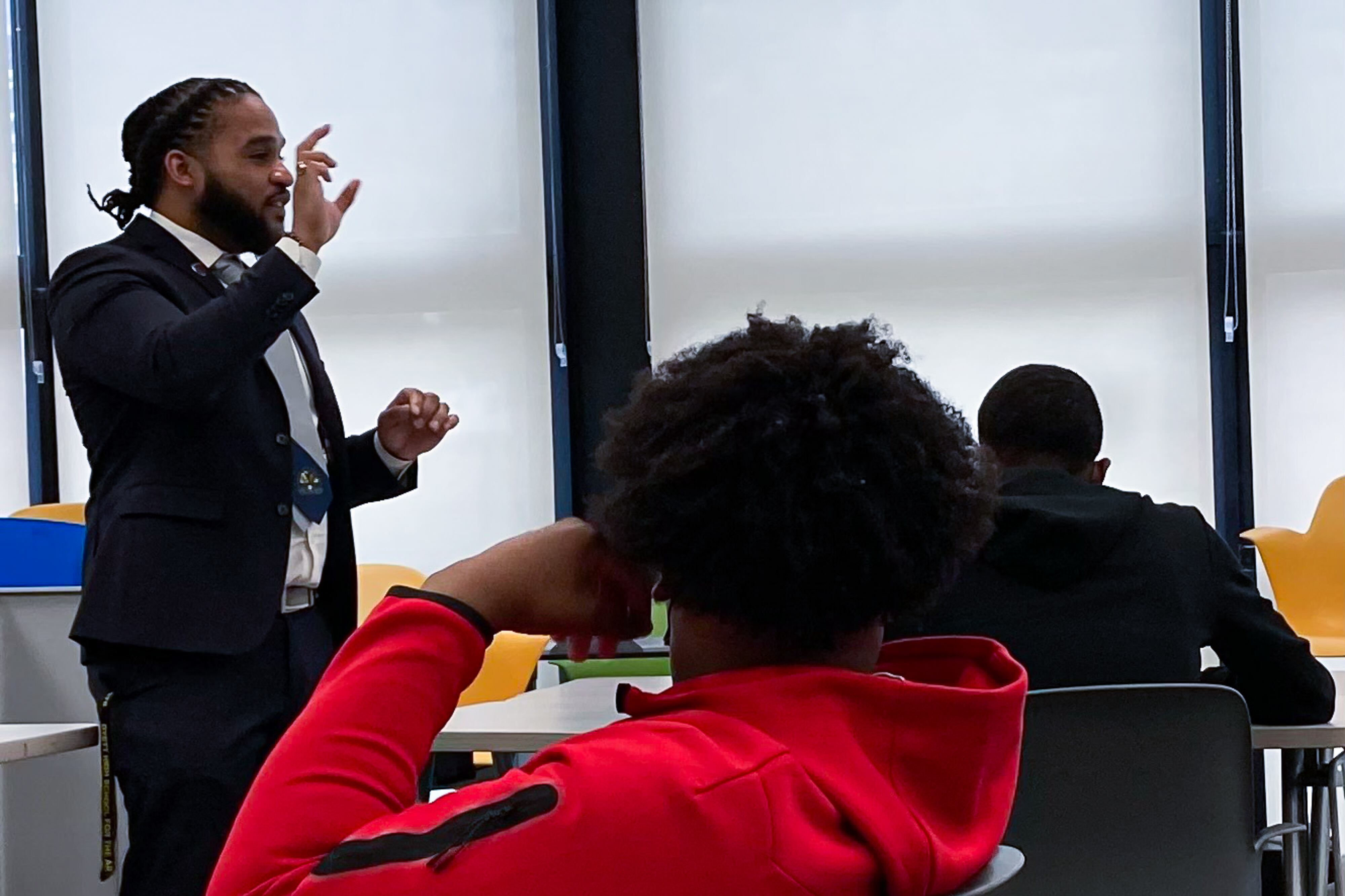 A man wearing a suit addresses two young men sitting at desks.
