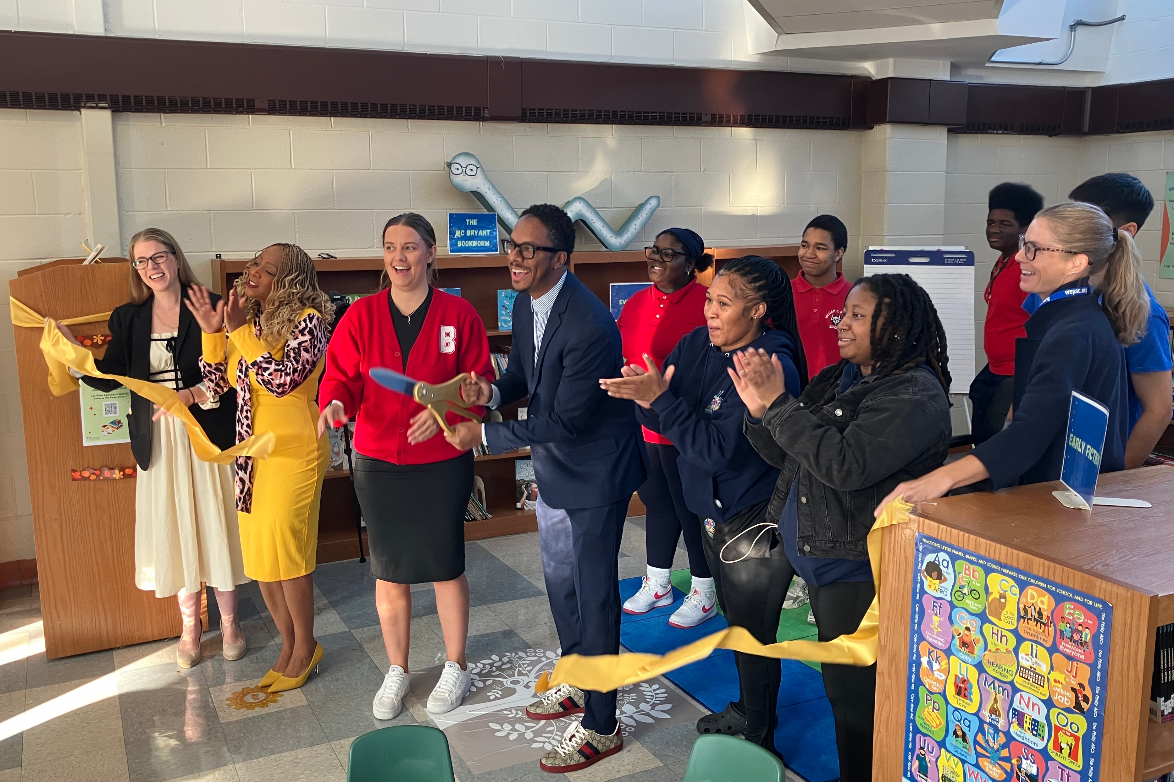 A photograph of a group of adults celebrating after cutting a yellow ribbon in a school library.