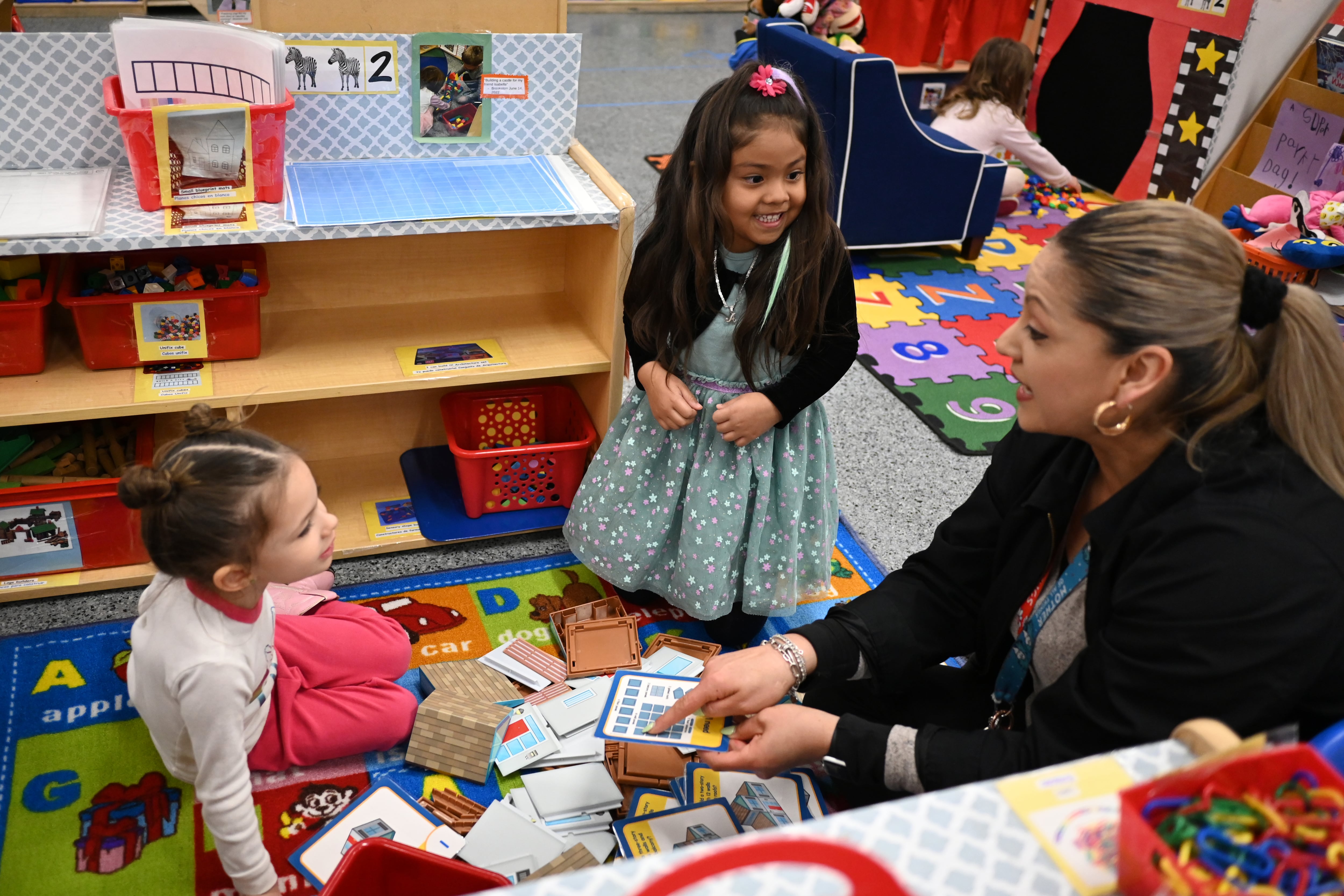 A teacher wearing gold hoop earrings sits on the floor of a classroom, over a rug with the alphabet, and points at pictures on cards while talking to two young girls.