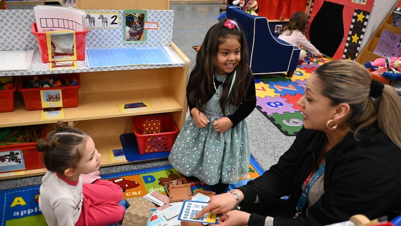 A teacher wearing gold hoop earrings sits on the floor of a classroom, over a rug with the alphabet, and points at pictures on cards while talking to two young girls.