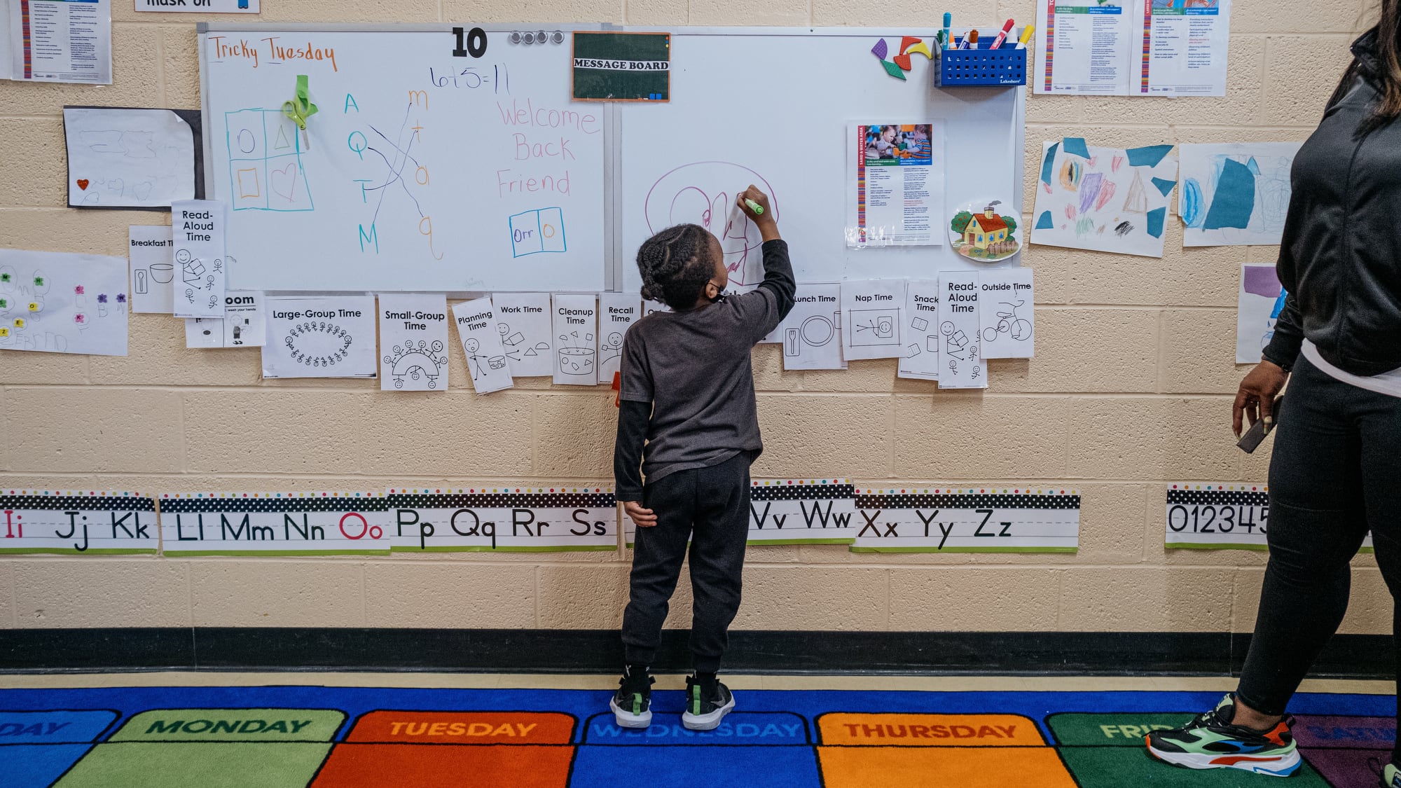 A preschool-age student writes on a white board in a school classroom.