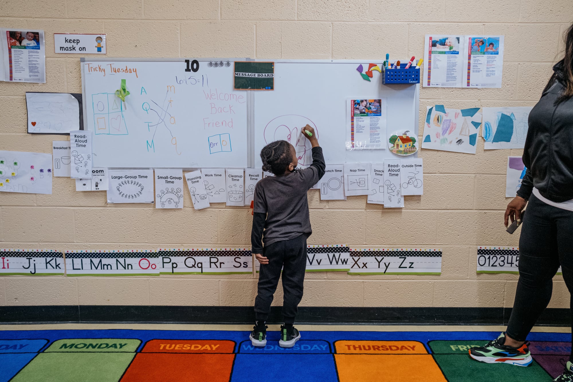 A preschool-age student writes on a white board in a school classroom.