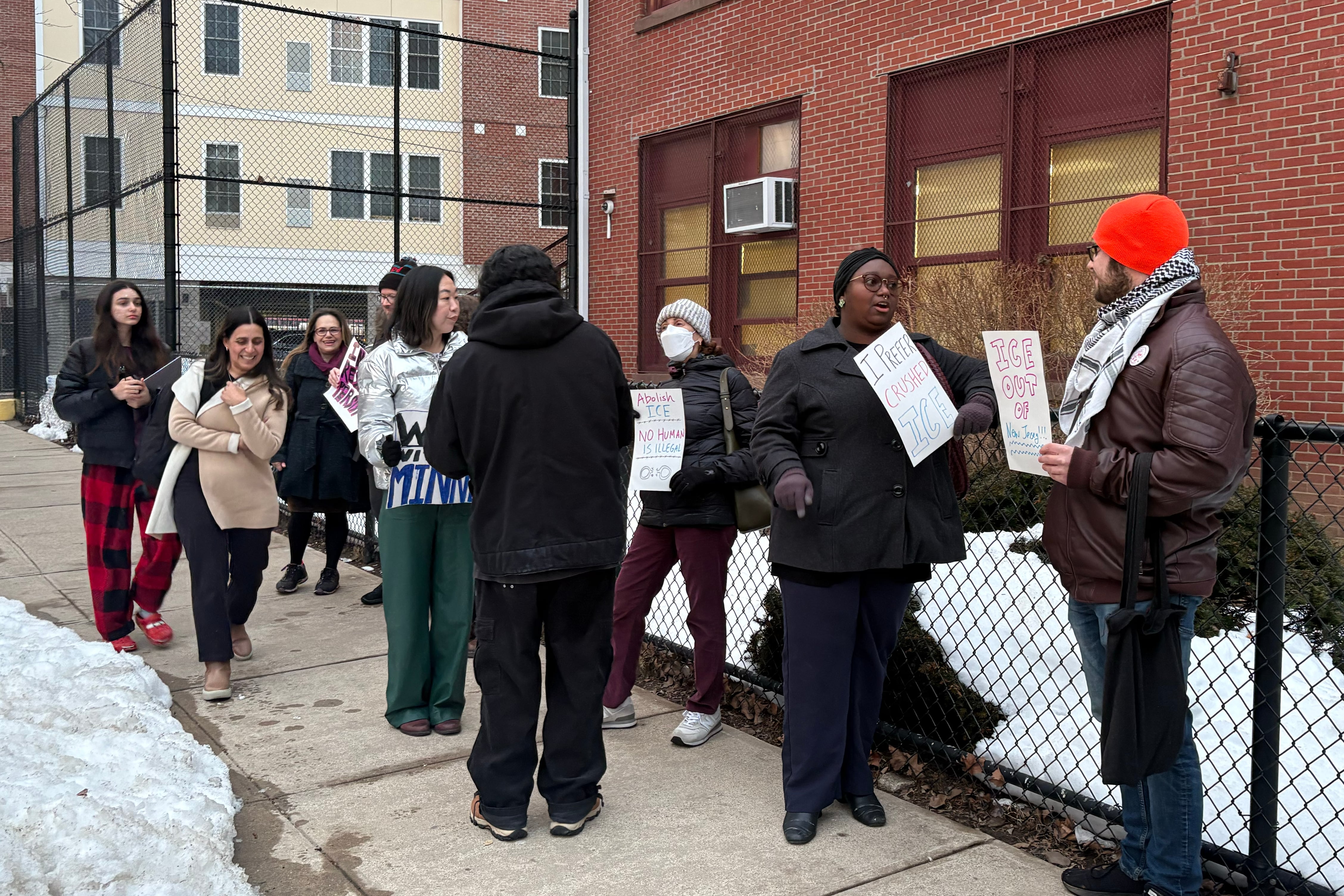 A photograph of people in winter jackets and hats some are holding protest signs on a cloudy day.