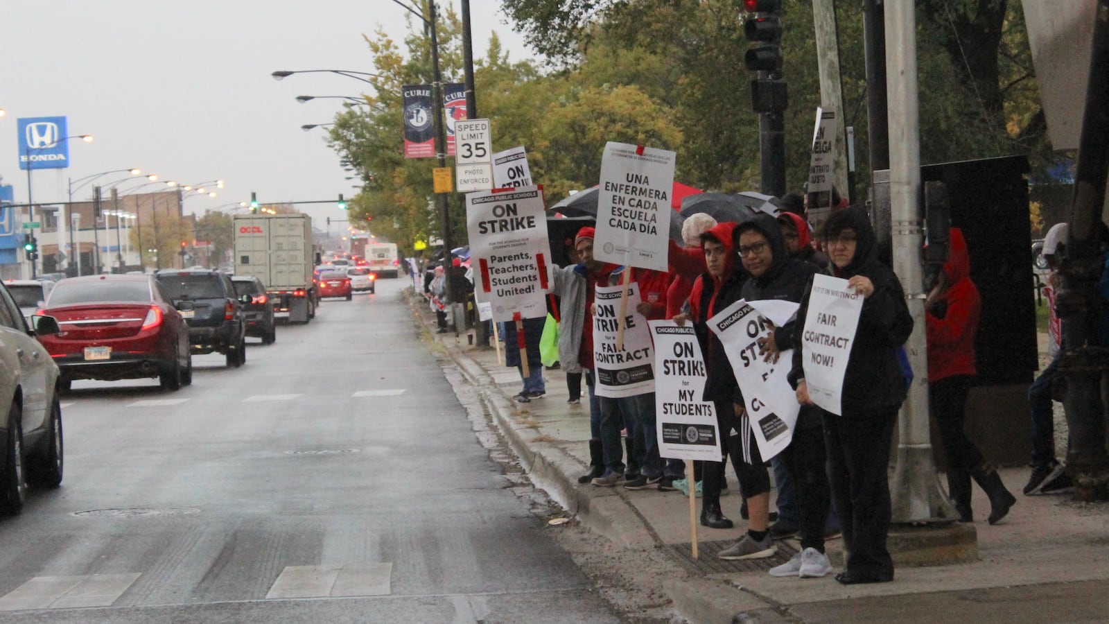 Striking Chicago teachers hold signs in front of Curie Metropolitan High School at the start of a lengthy picket line down Pulaski Road. Rain in the forecast leads some passersby to drop off ponchos.