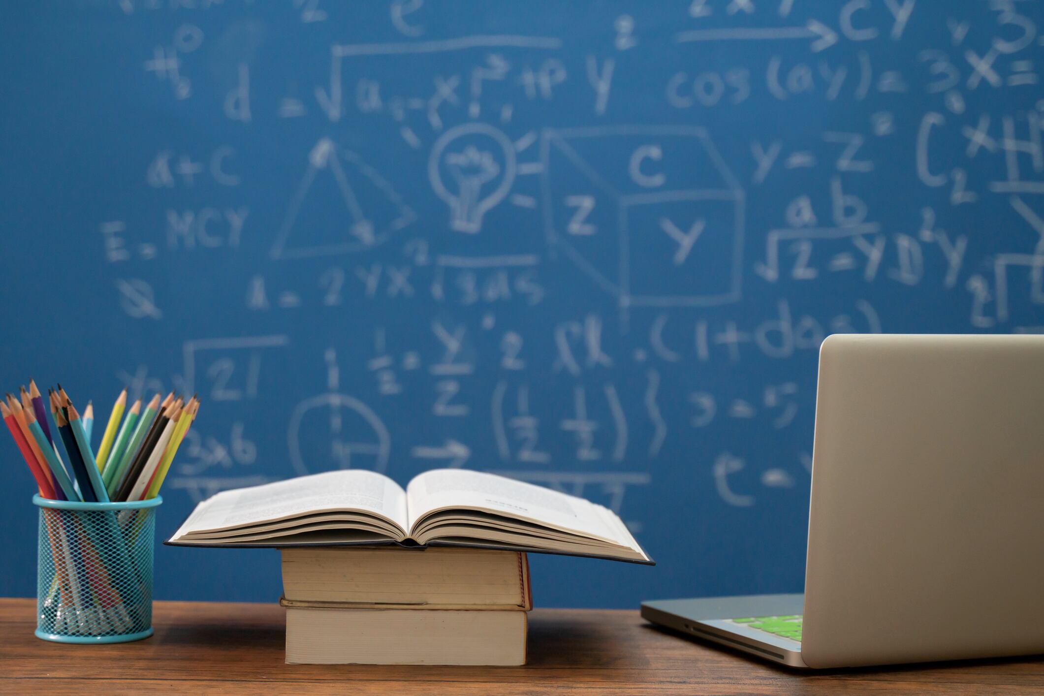 Books, colored pencils, and a laptop on a wooden table with a blackboard in the background.