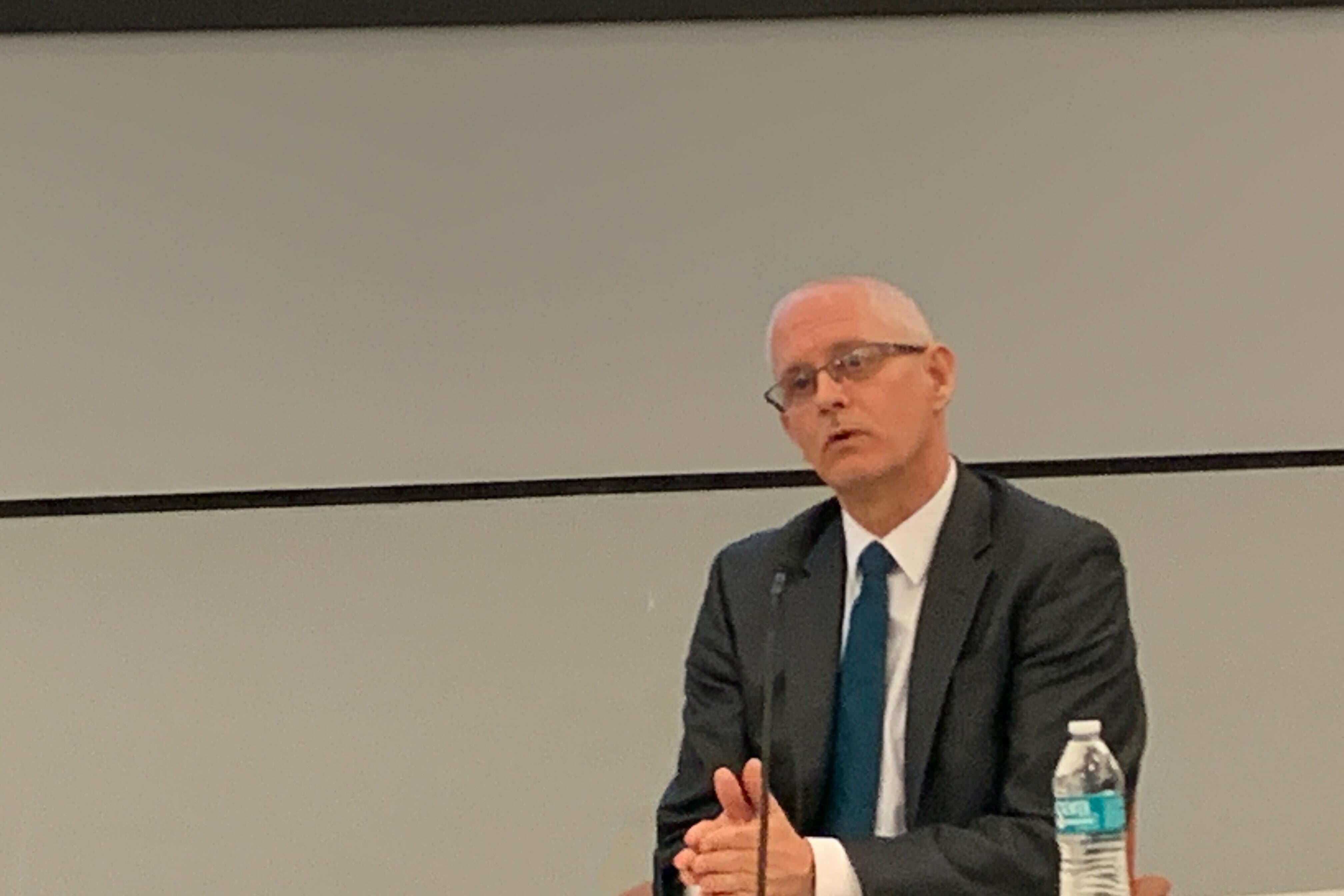 John L. Davis speaks into a microphone while sitting at a table. He is wearing a dark suit and blue tie, and there is a water bottle in front of him.