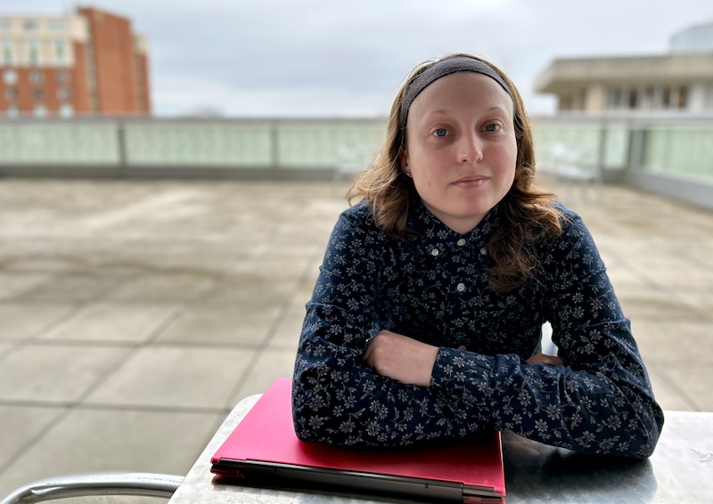 A young woman with short blonde hair sits at a table with her books outside. There is grey stone floor and a cloudy sky in the background.