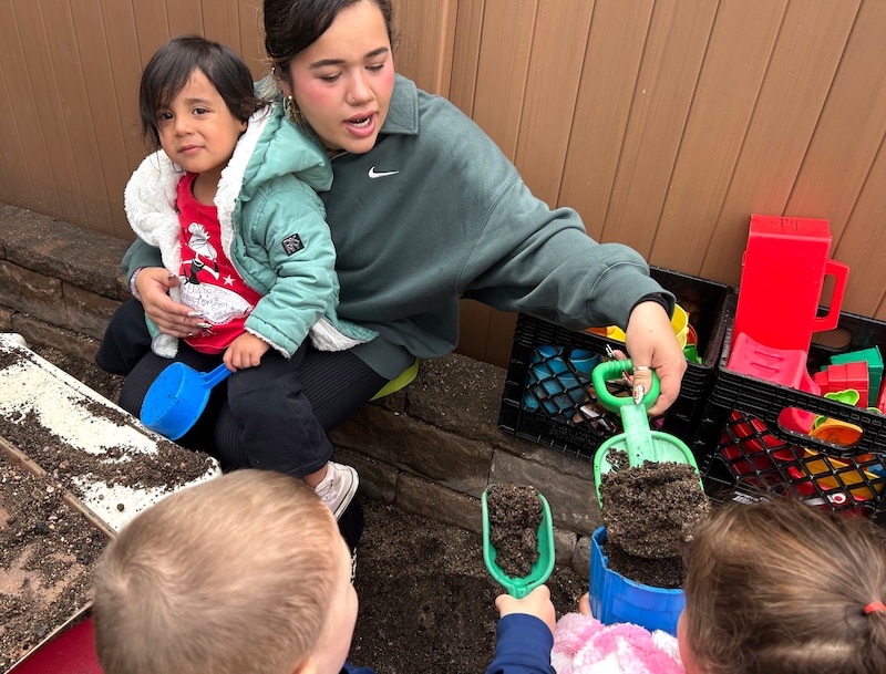 A woman with a green top holds a child in her lap and with the other hand uses a little green shovel.