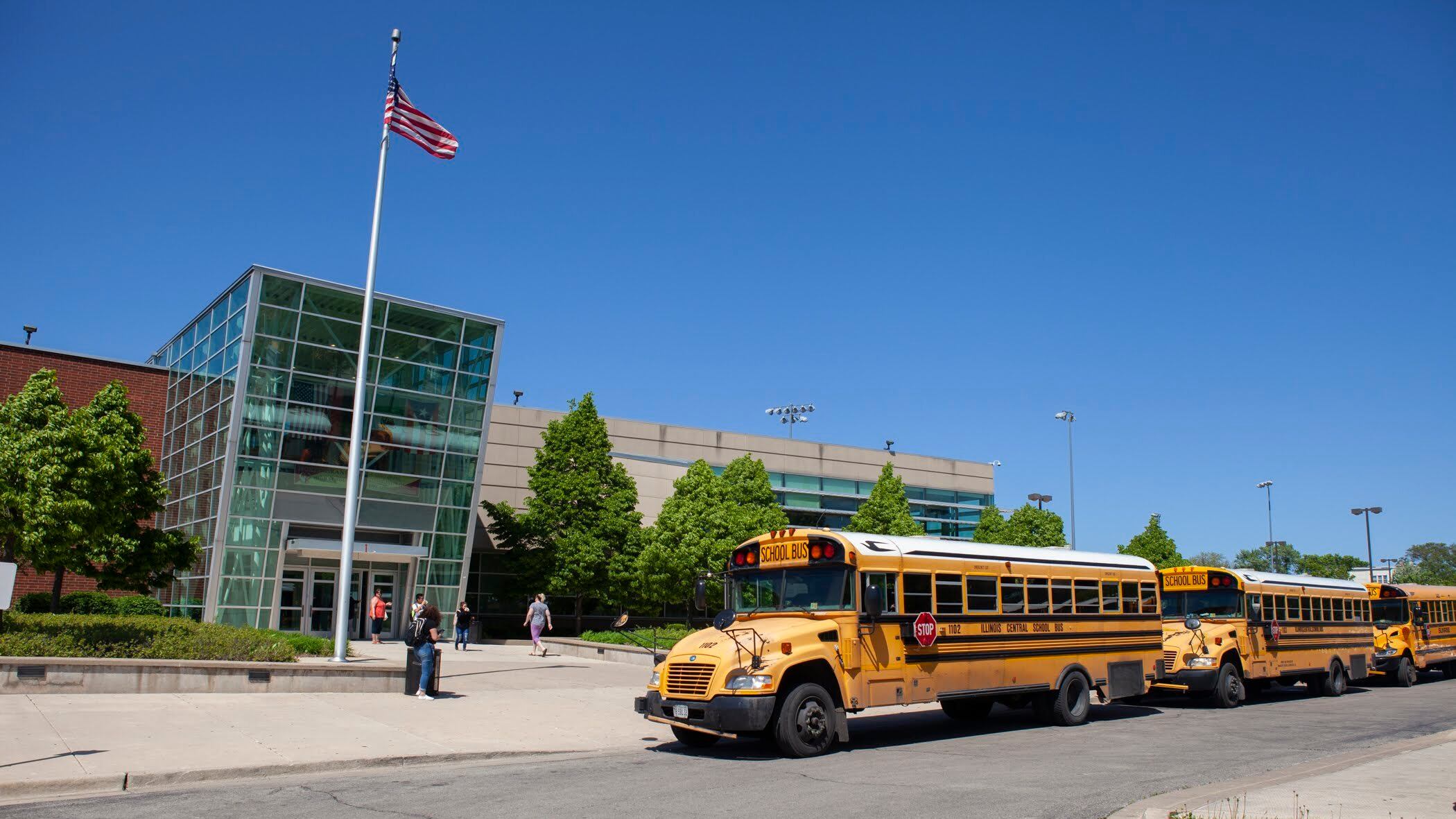 School buses and an American flag are at the front of North-Grand High School in Chicago, with school buses lined up in front of the campus.