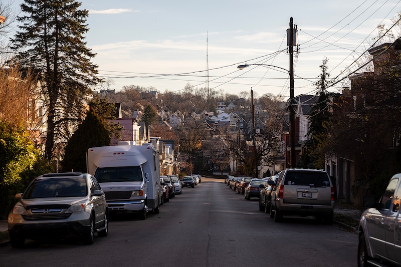 Parked cars and street lights line each side of the street with a hill full of houses in the background.