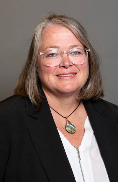 A woman with medium length hair, wearing glasses and a dark suit jacket poses for a portrait in front of a grey background.