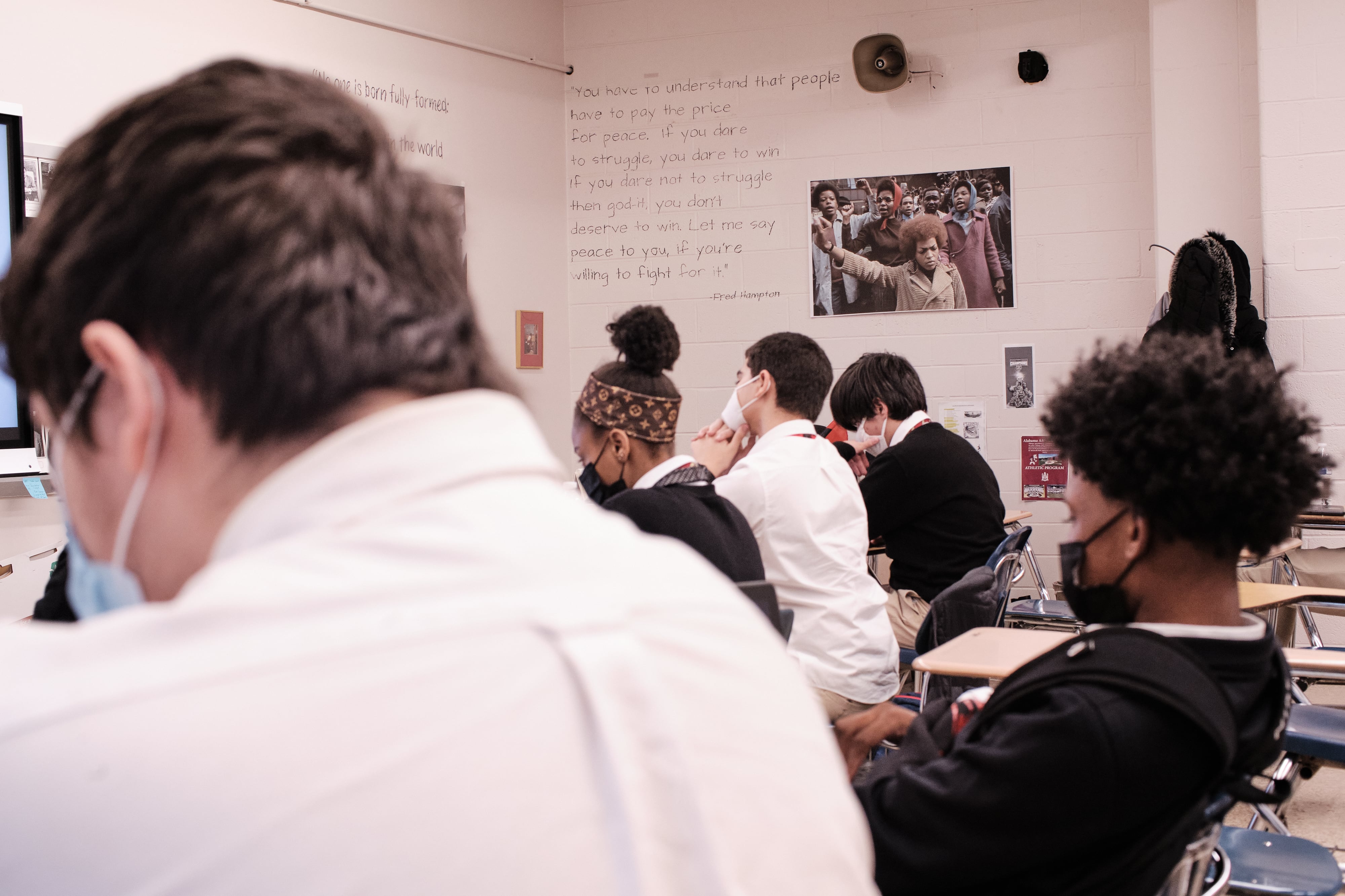 Students sit in a classroom wearing different color masks. On the wall to the right, there is a photo of a Black person rising a fist towards the camera.