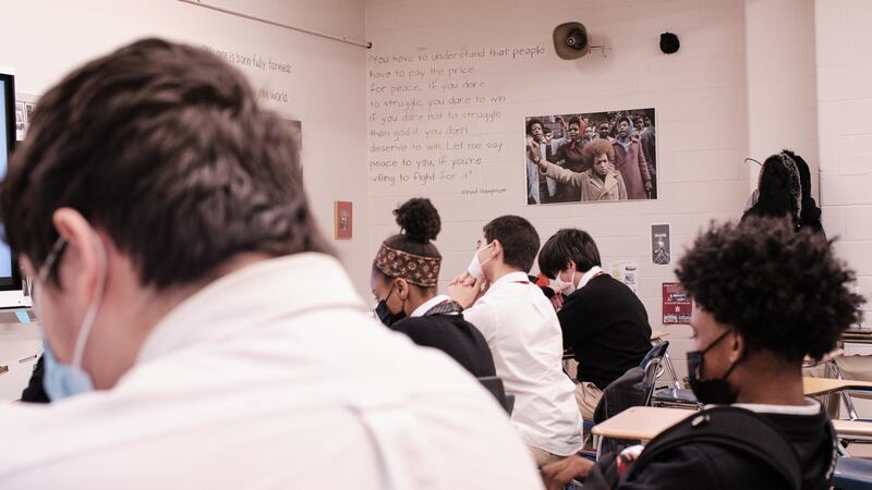 Students sit in a classroom wearing different color masks. On the wall to the right, there is a photo of a Black person rising a fist towards the camera.