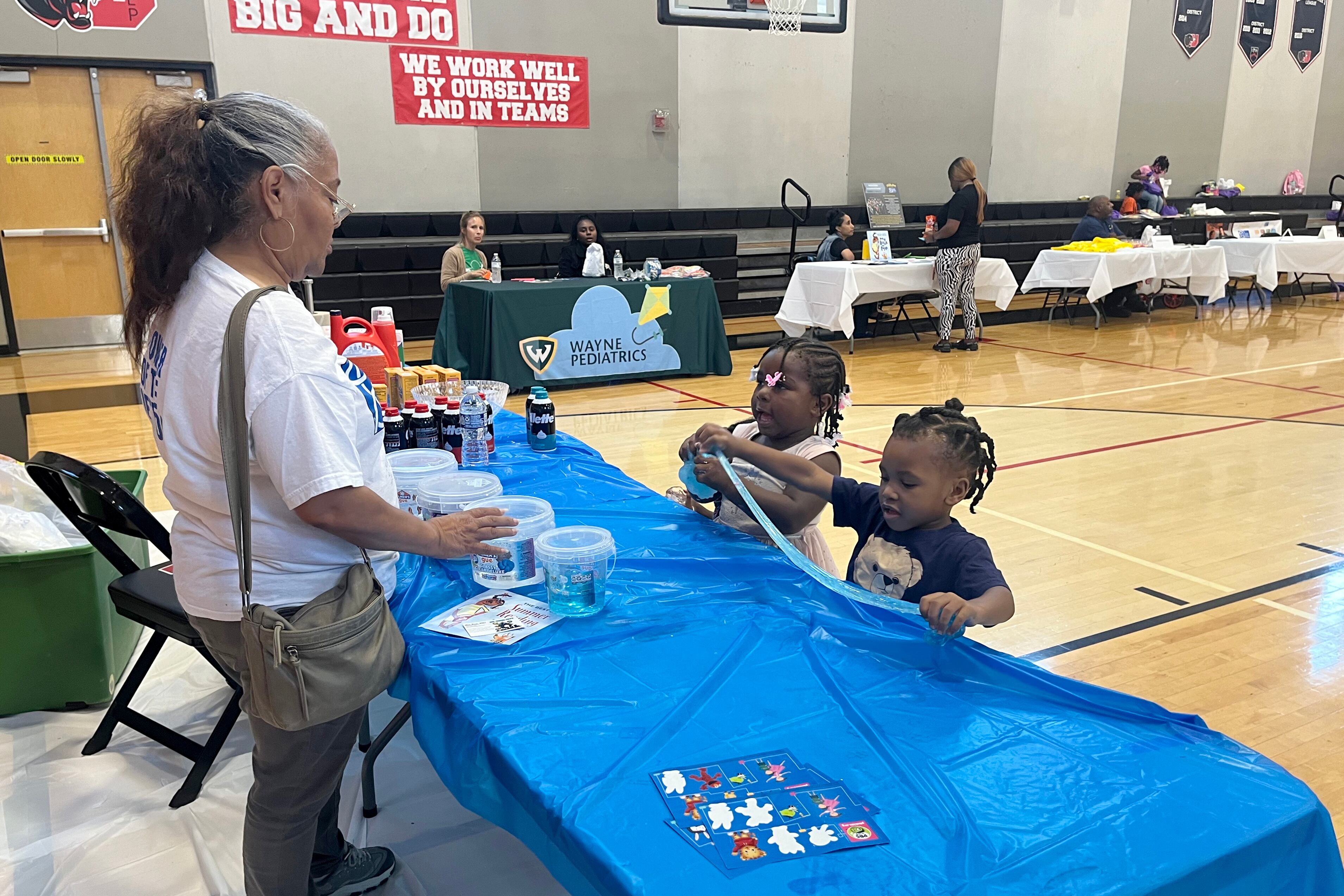 An adult and two young children stand at a table covered in a blue table cloth in a school gym.