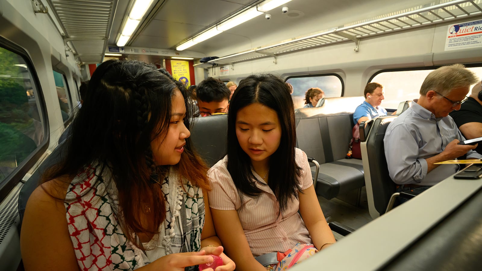 A photograph of two high school students sitting next to each other in a SEPTA train car. There are a few people sitting in different seats on the right of the students.