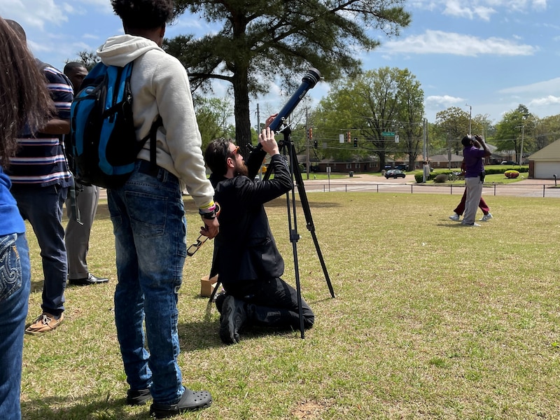 An adult looks through a telescope while a couple of students stand nearby with green grass and a clear blue sky.