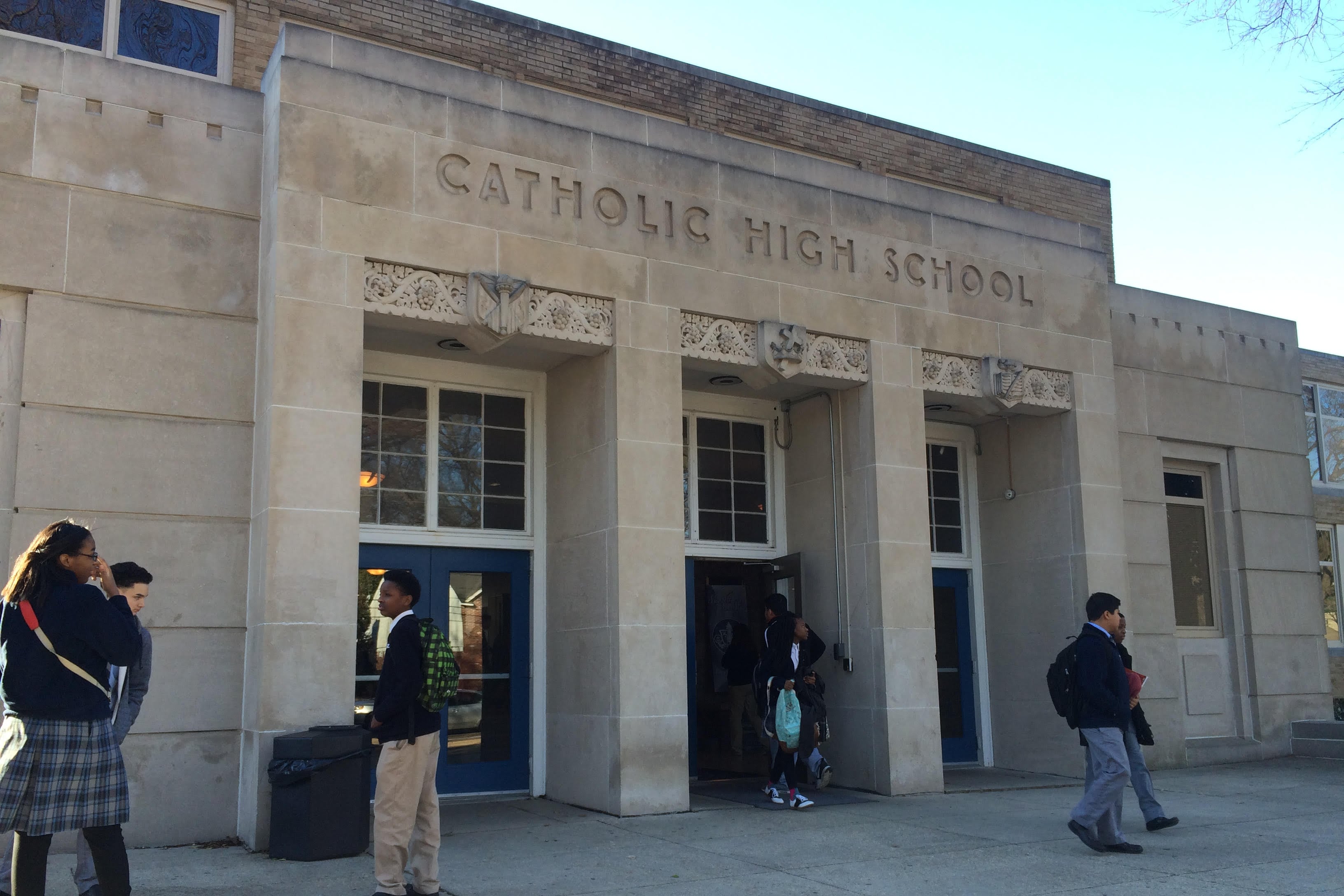 A photograph of high school students in uniforms stand outside of a large stone building with words at the top of the building that reads "Catholic High School."