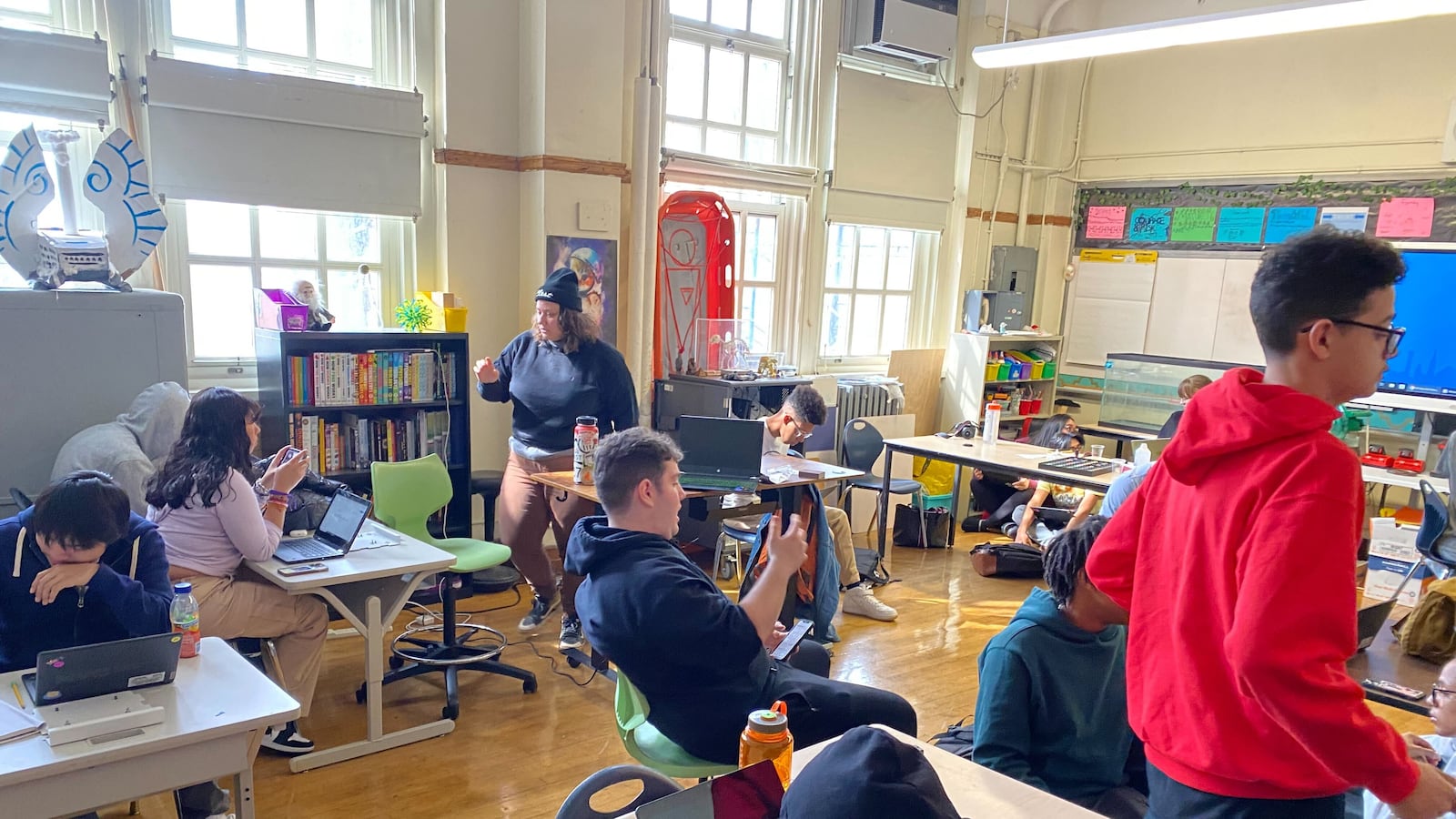 Teens sit at tables scattered around a classroom working on different projects.