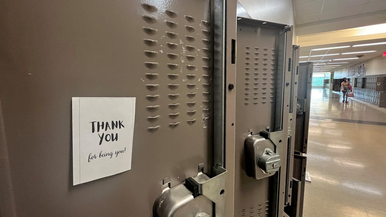 A “Thank You” note on white paper hangs on the inside of a light brown high school locker in the foreground with several lockers in the background while a student walks down the hallway.