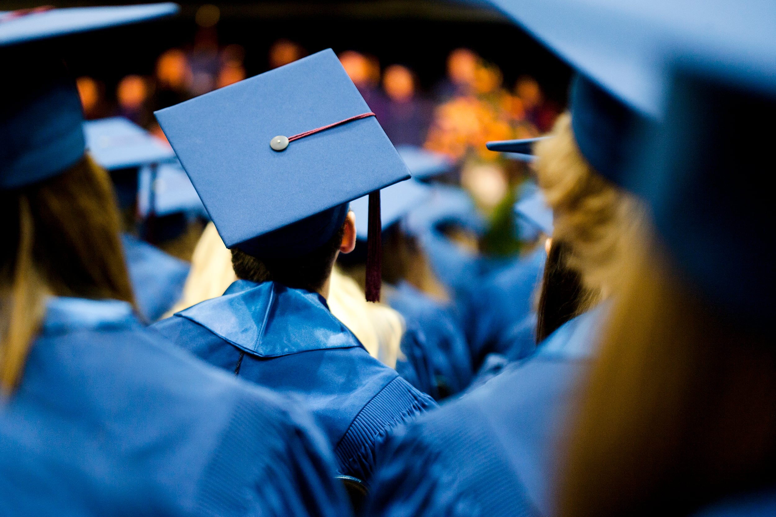 A photograph of a high school student wearing a blue graduation cap and gown surrounded by a sea of high schoolers in graduation gowns.