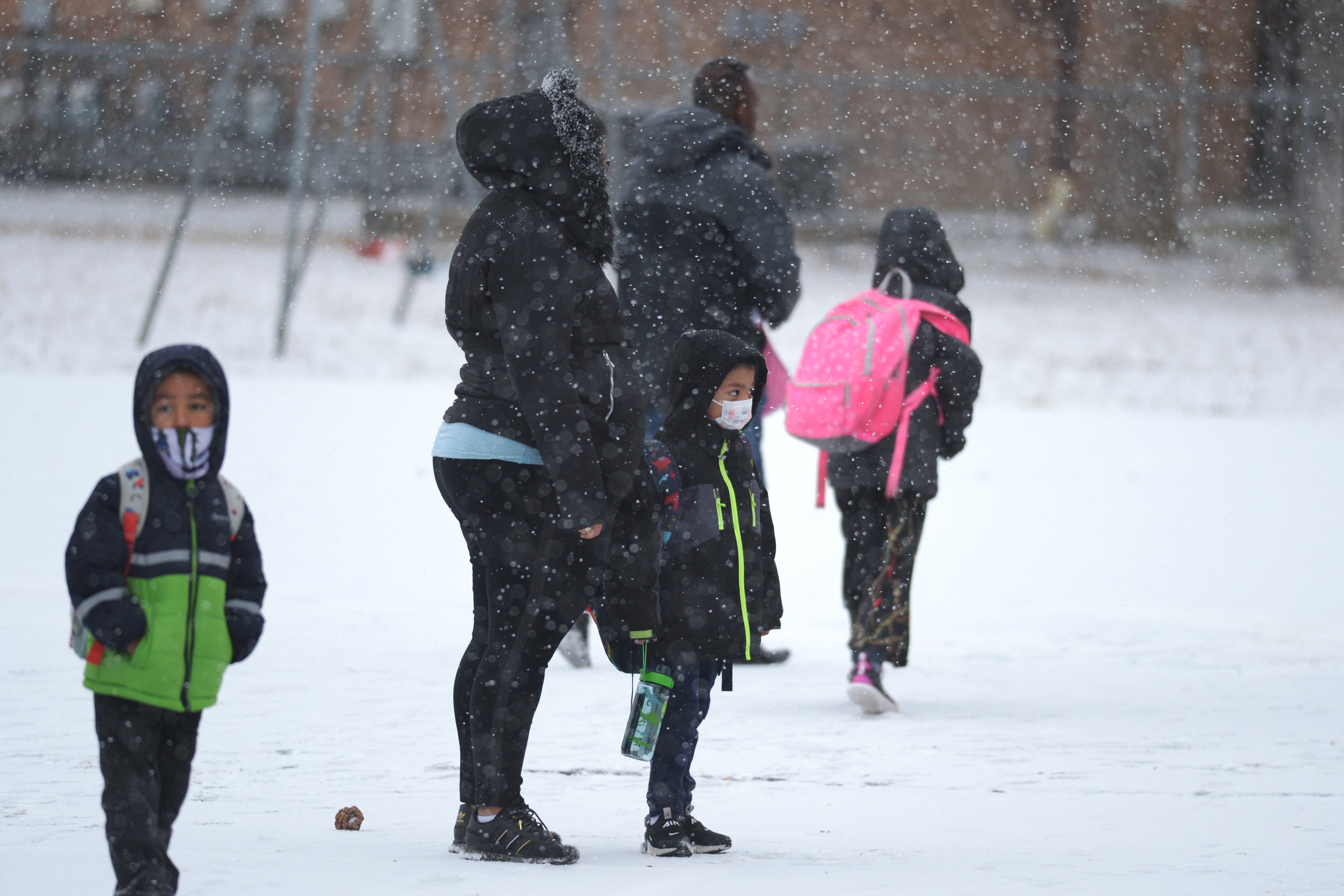 Parents and young children wearing masks and winter coats wait outside a school building on a snowy day.