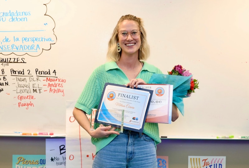 A woman in a green shirt and jeans holds a certificate and a bouquet of flowers.