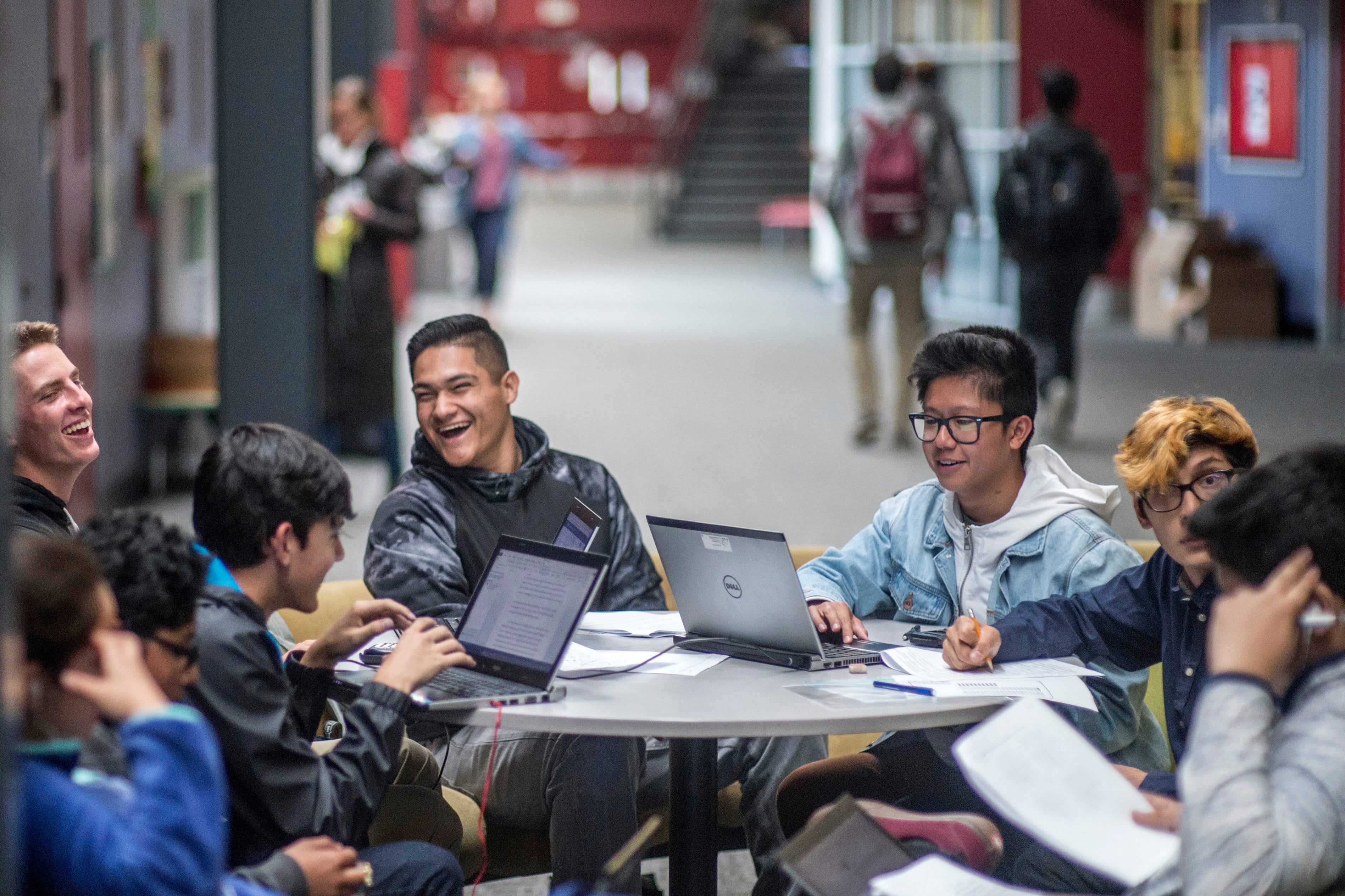 A group of high school students laugh and talk while sitting around a table.