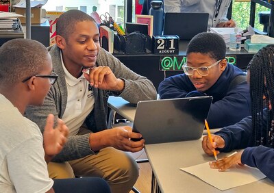 An adult kneels next to a desk with three students working.