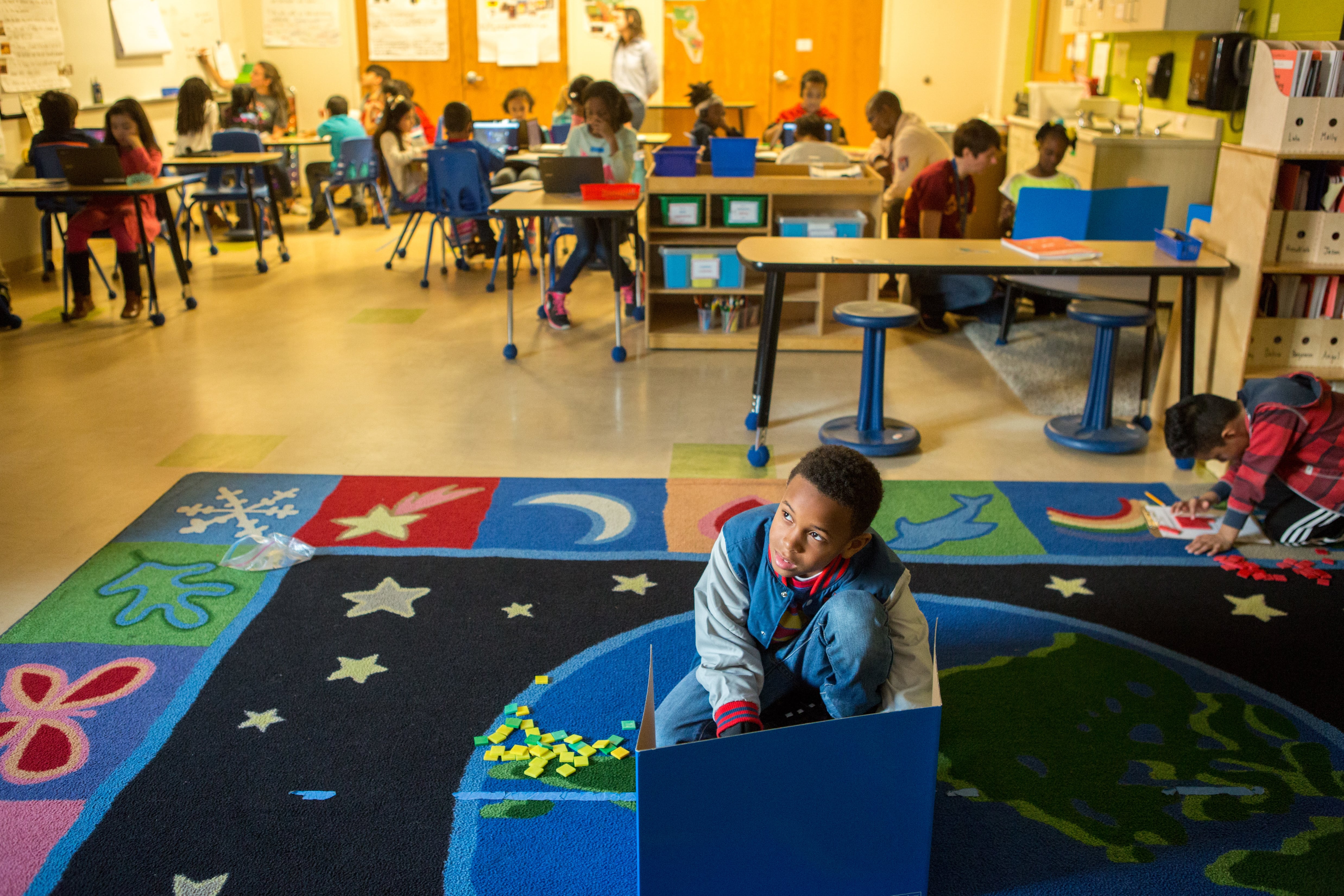 A young students sits on a colorful rug working on a project while the rest of the class is working from desks in the background.
