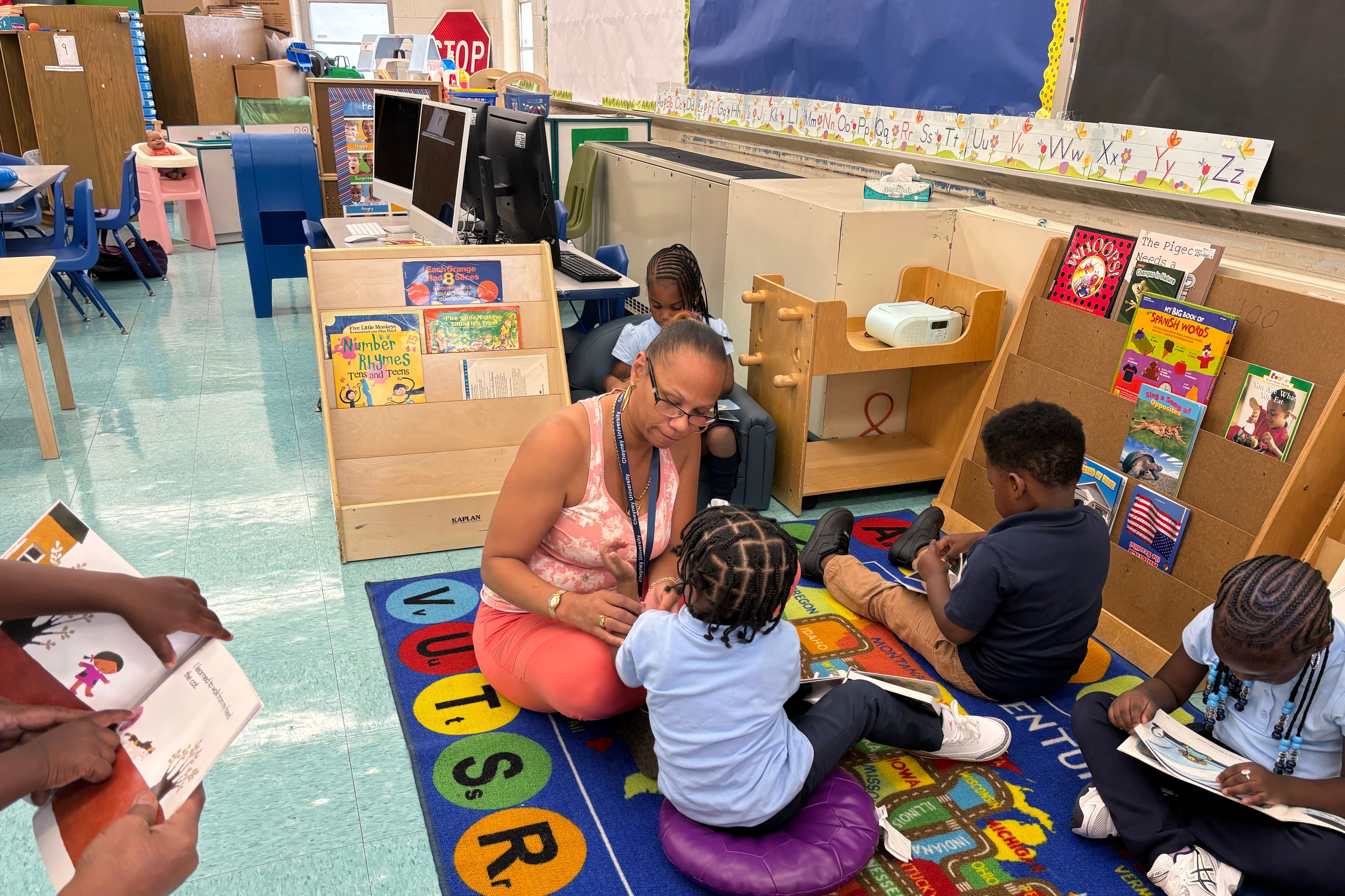 An adult wearing a pink top sits with a group of young children on a colorful rug in a classroom.