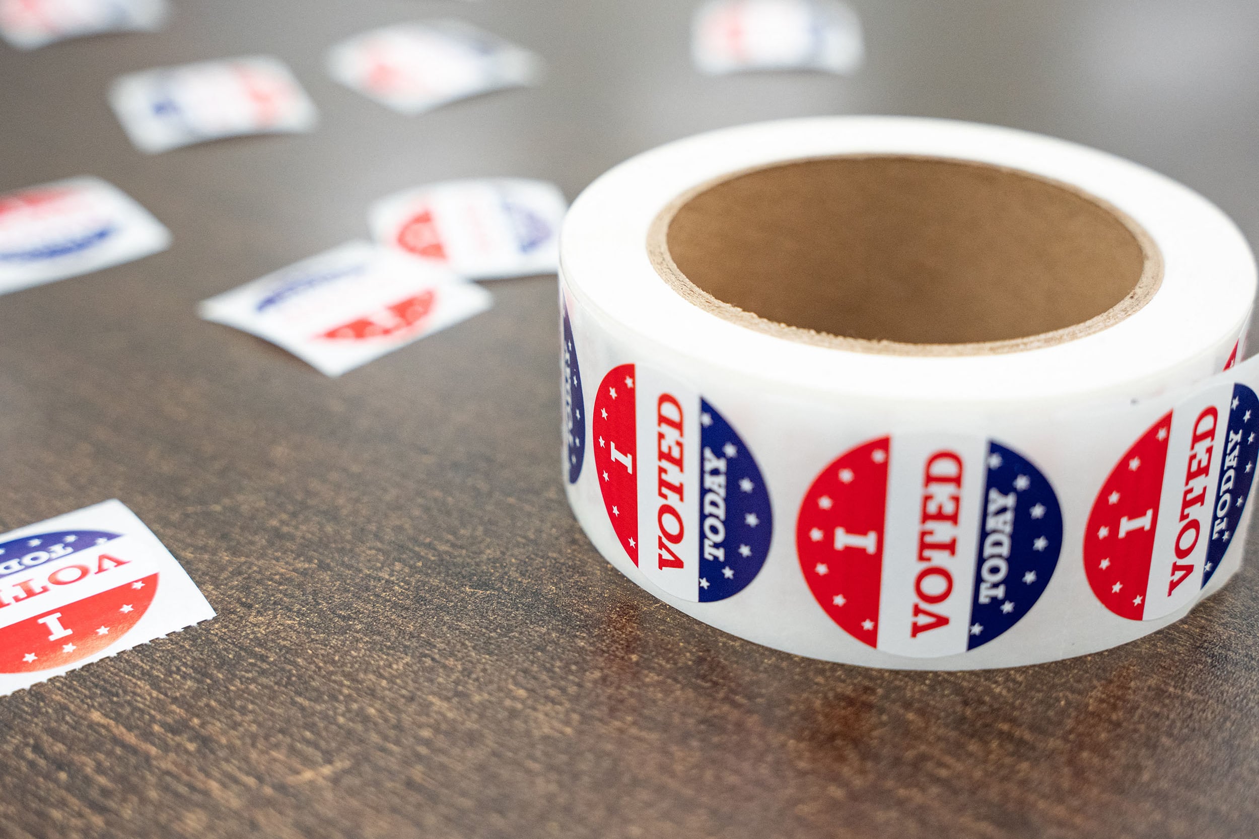 A roll of red, white and blue "I voted today" stickers sit on a wooden table with other individual stickers around the roll.