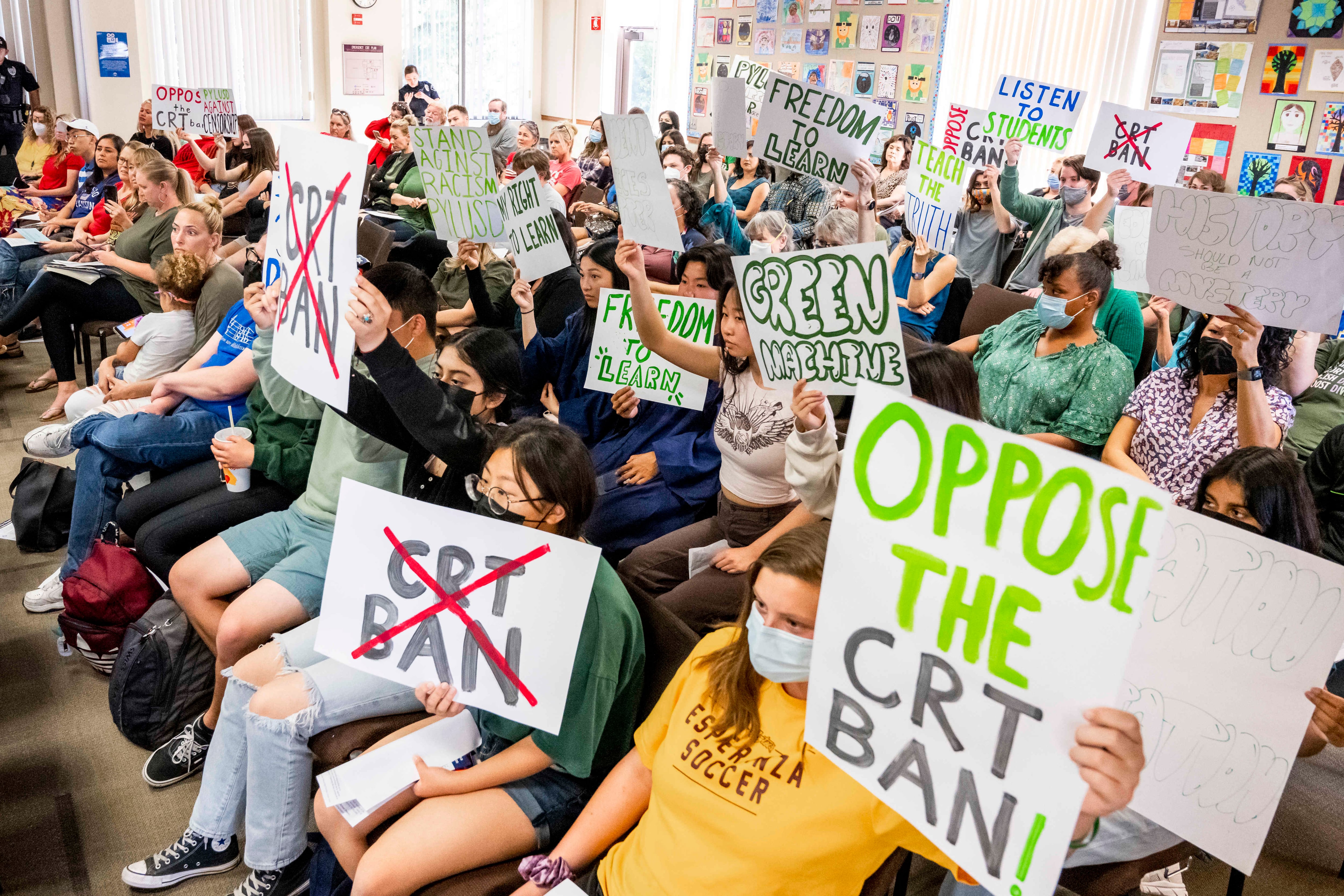 Masked students sitting in chairs hold up signs opposing a proposed ban on teaching CRT, in a room of adults and students.