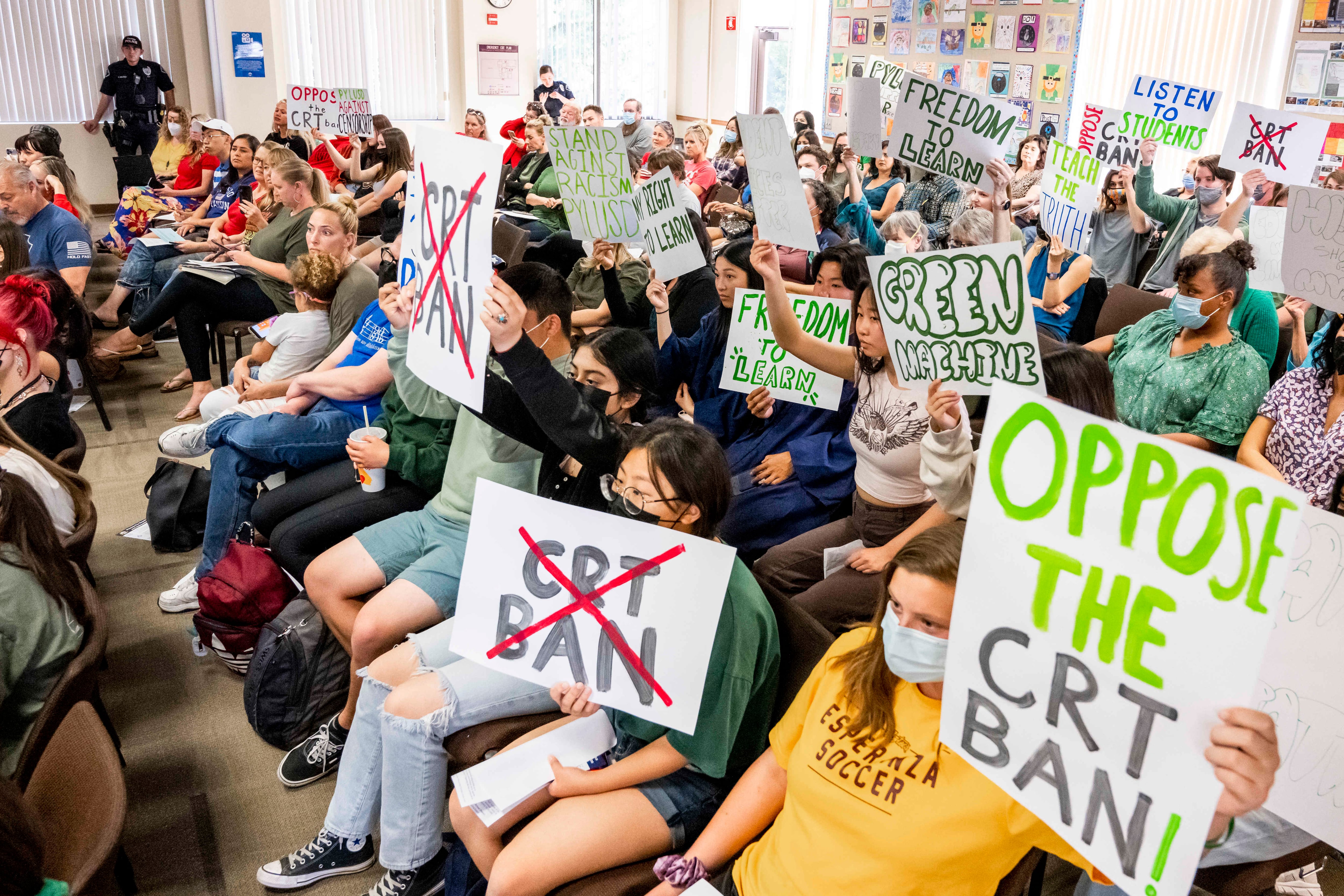 Masked students sitting in chairs hold up signs opposing a proposed ban on teaching CRT, in a room of adults and students.
