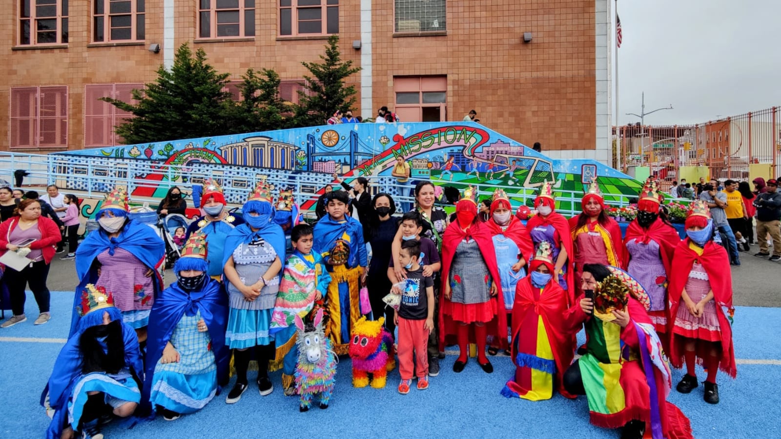 Parents and students in bright blue and red costumes line up in front of a school mural outside of a brick building.