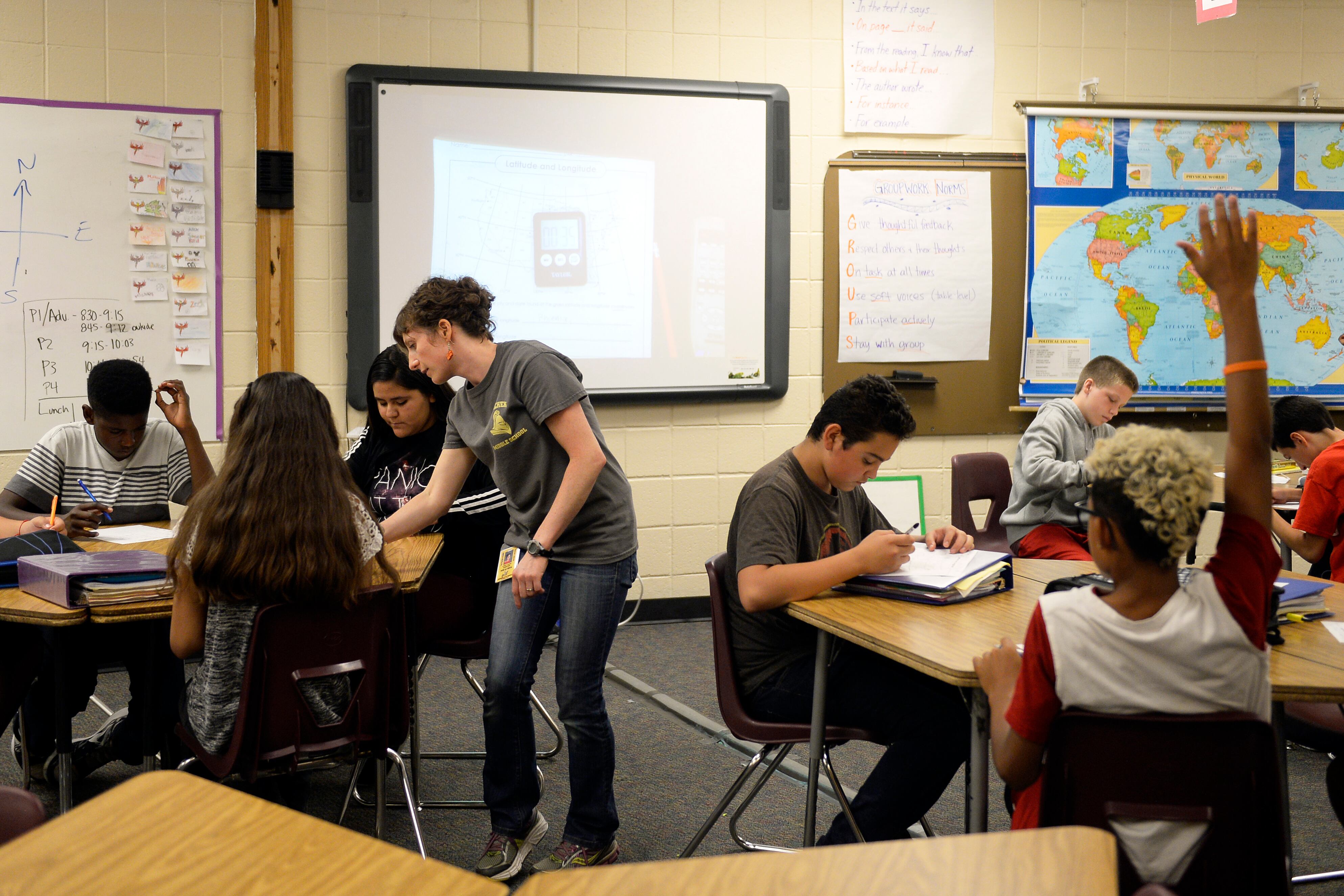 A teacher wearing a grey shirt talks with one of several students sitting at a desk in a classroom. There is a whiteboard, projected screen and a large world map on the wall in the background.