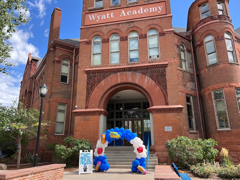 A balloon arch with a welcome sign in front of the front entrance to a red brick school building with the words "Wyatt Academy" on the top of the building.