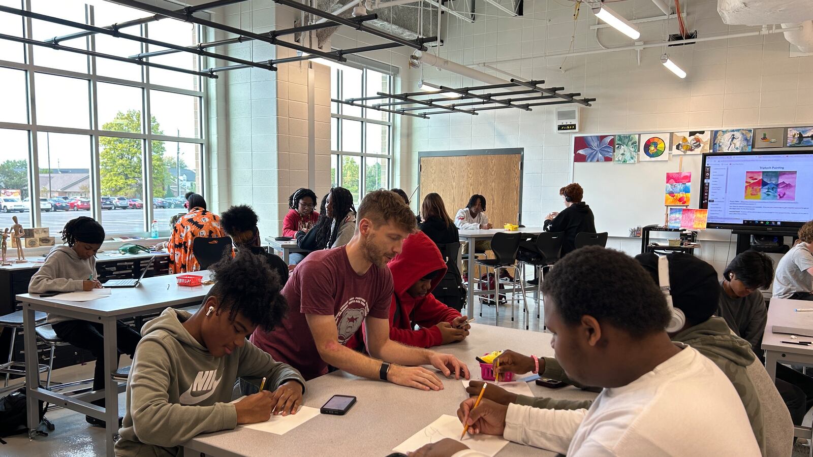 Students sit at desks in a bright classroom with high windows, working on art projects.