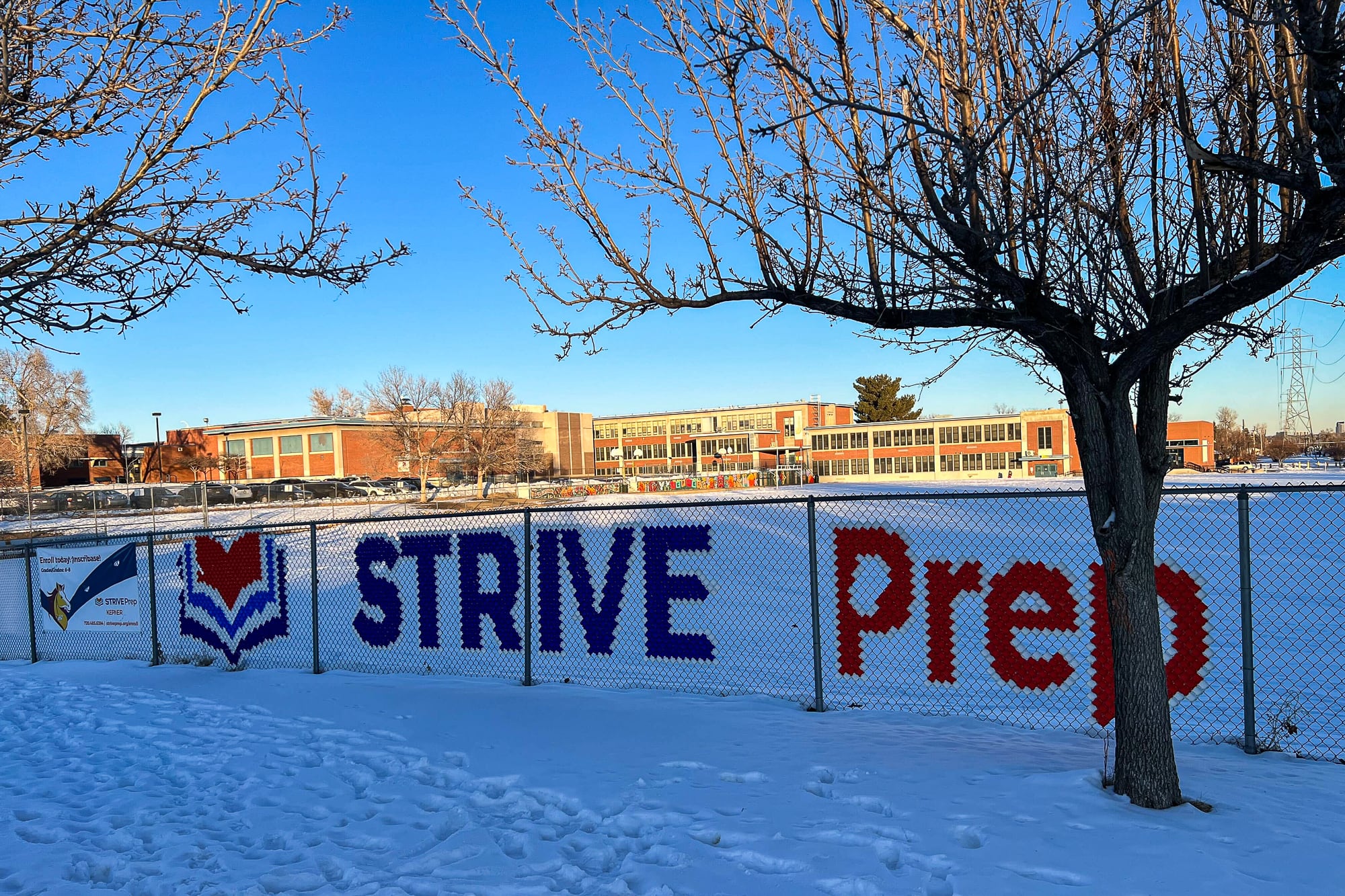 A fence that says “STRIVE Prep” sits in front of a school building. Snow covers the ground.