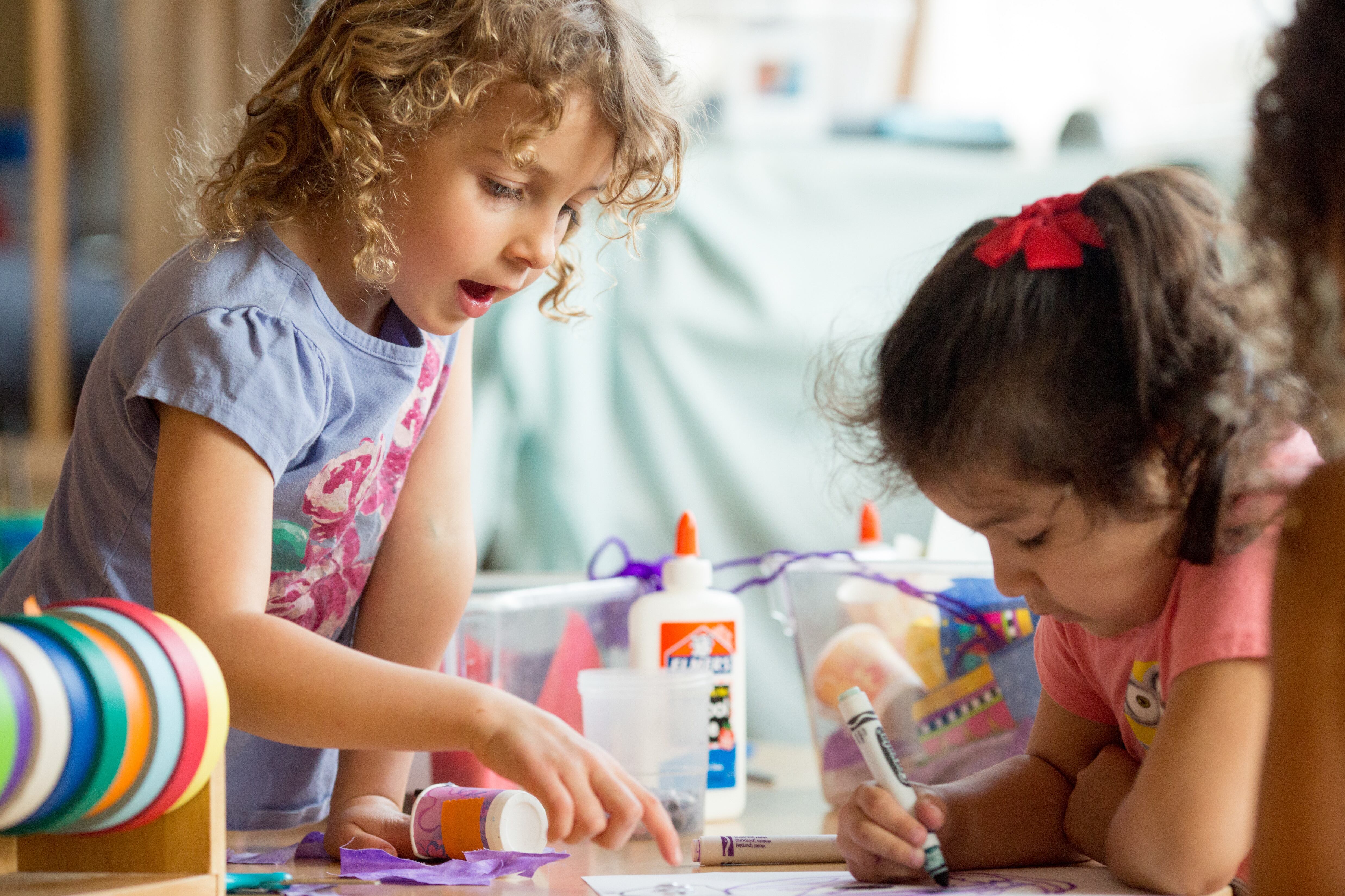 A preschool girl points to something on a table, while another girl works nearby.