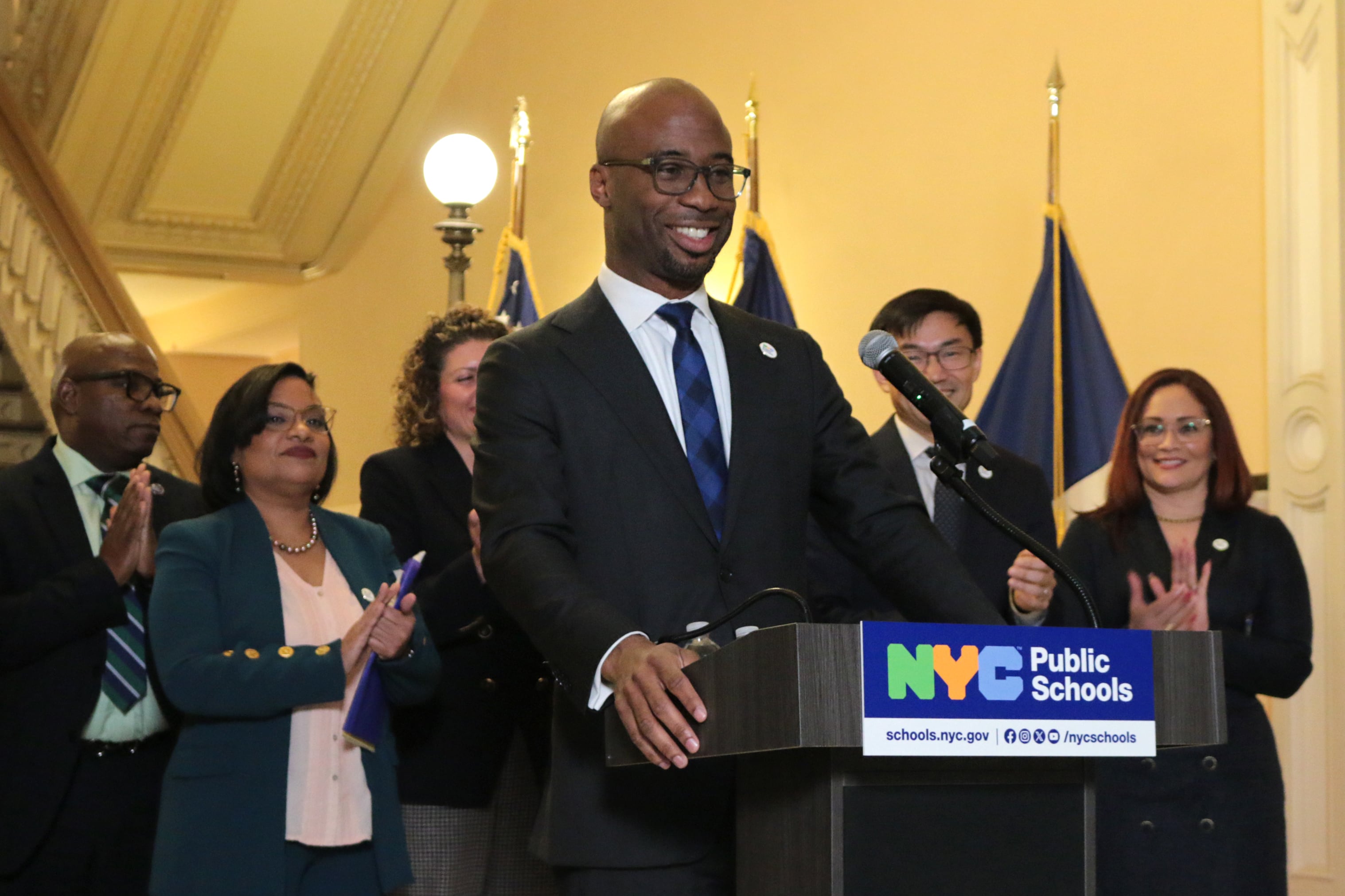 A photograph of a Black man in a suit standing behind a podium with people standing behind him.