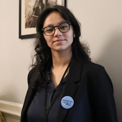 Headshot of a teenage girl with black hair. She wears a black top with a blue button that says "Journalism for All."