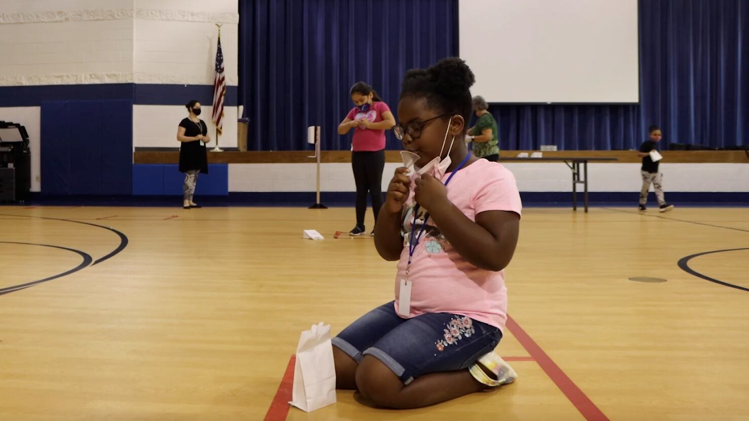 A young girl produces a saliva sample for a SHIELD COVID test inside of a large gymnasium.