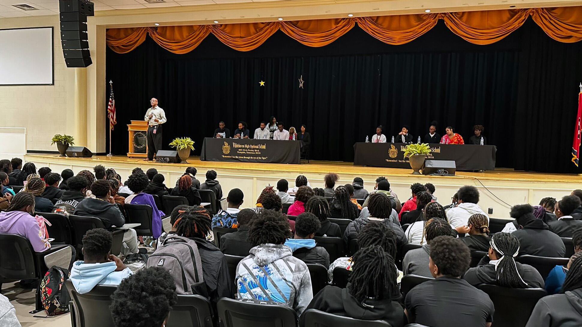 Several students are facing a stage. On stage are two small tables with speakers, and a podium with a speaker.