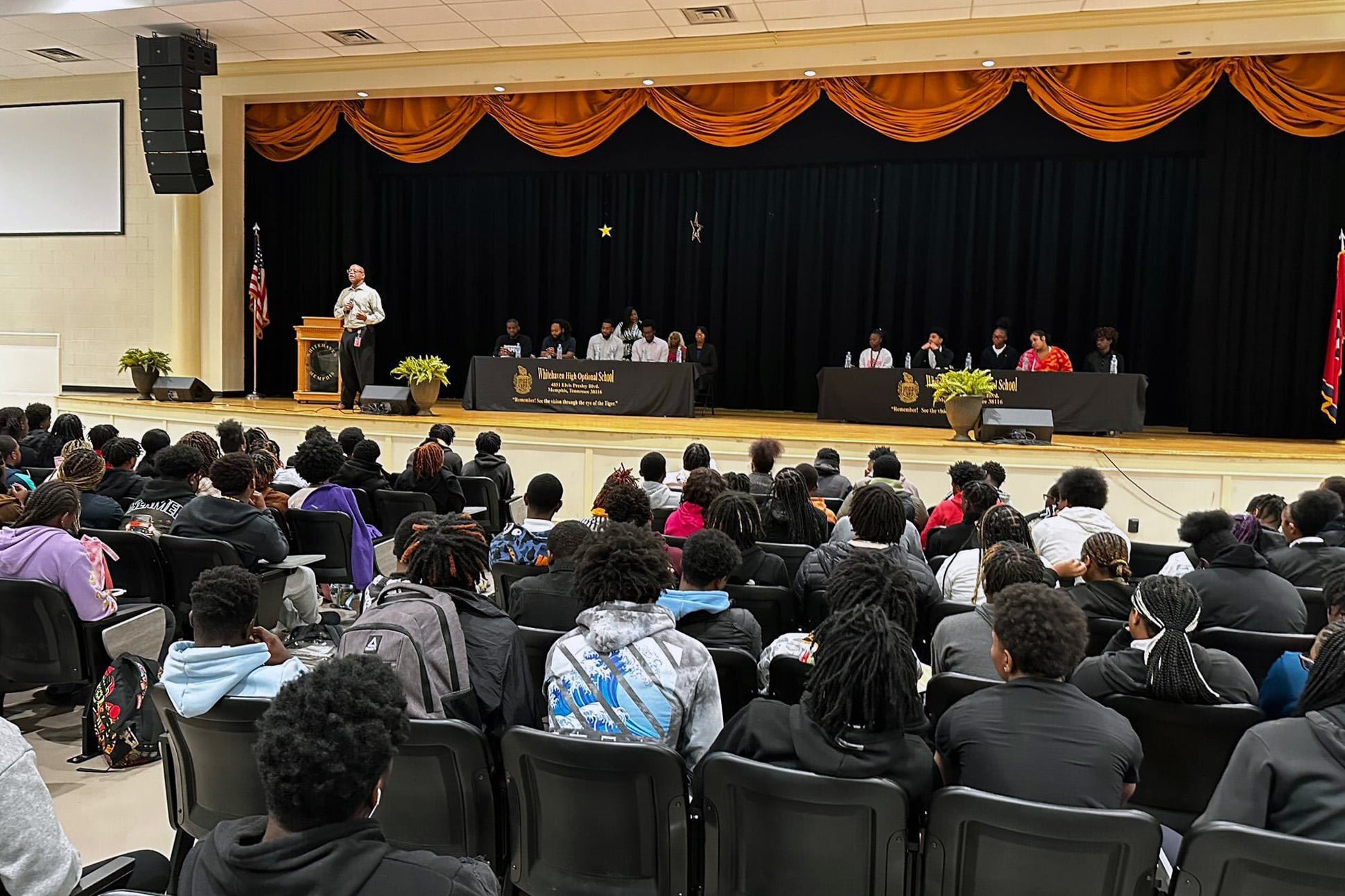 Several students are facing a stage. On stage are two small tables with speakers, and a podium with a speaker.