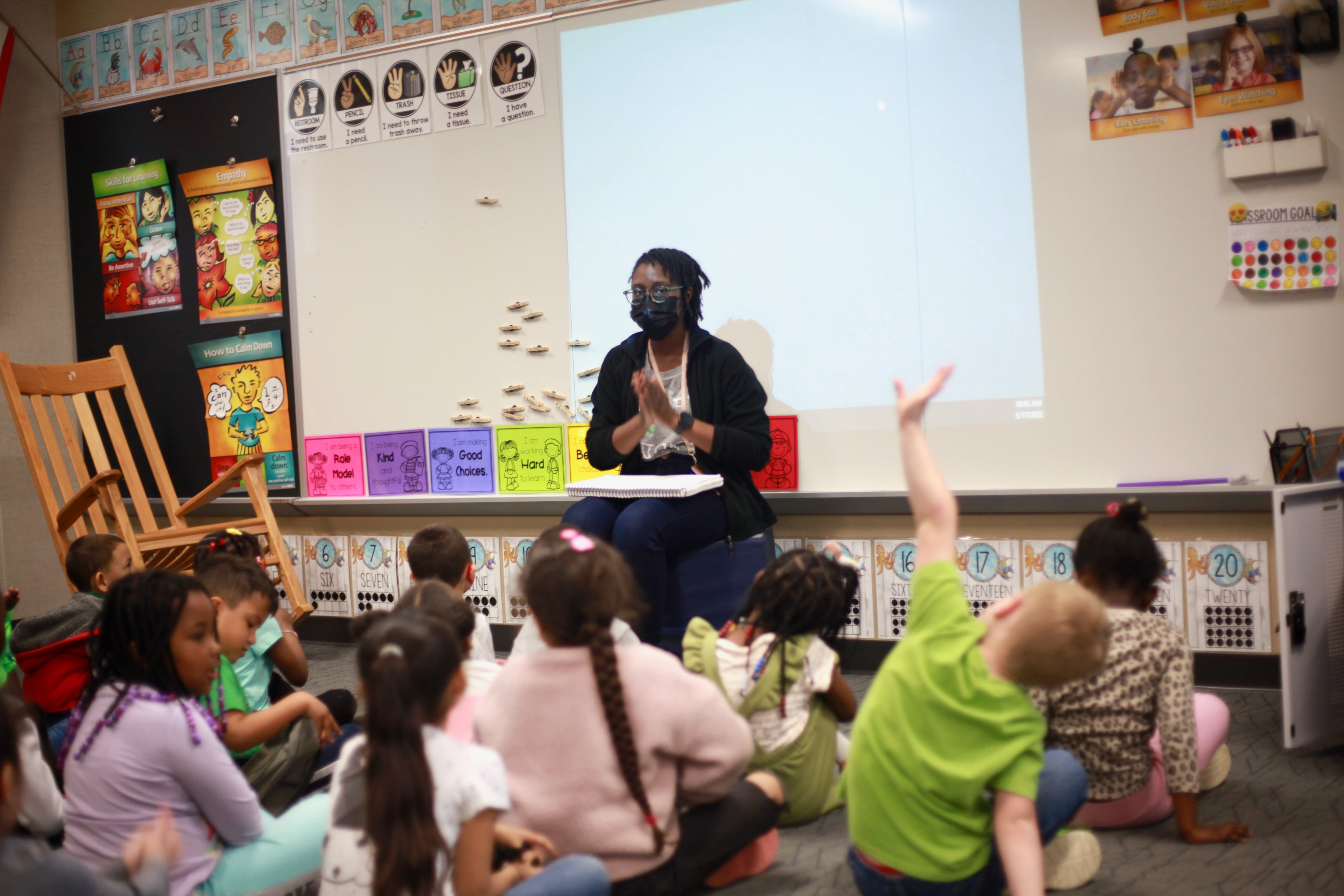 Students sit on a rug and raise their hands during class, as their teacher claps her hands together at the front of the class.